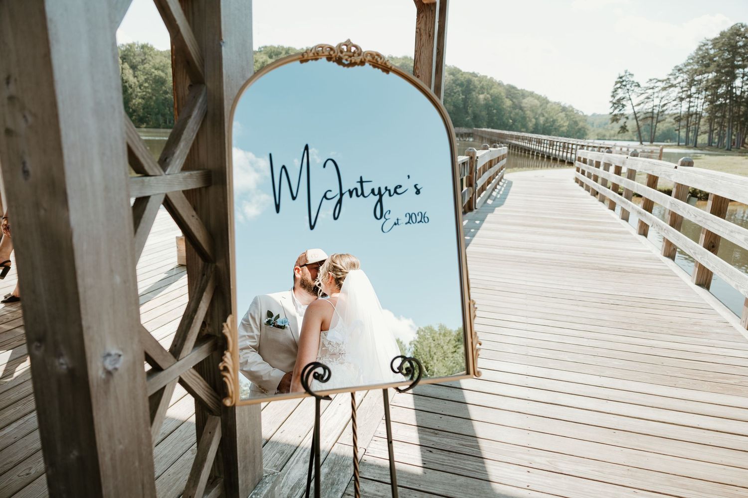 Newlyweds kiss reflected in ornate gold mirror displaying McIntyre's Est. 2024 sign on rustic wooden bridge.