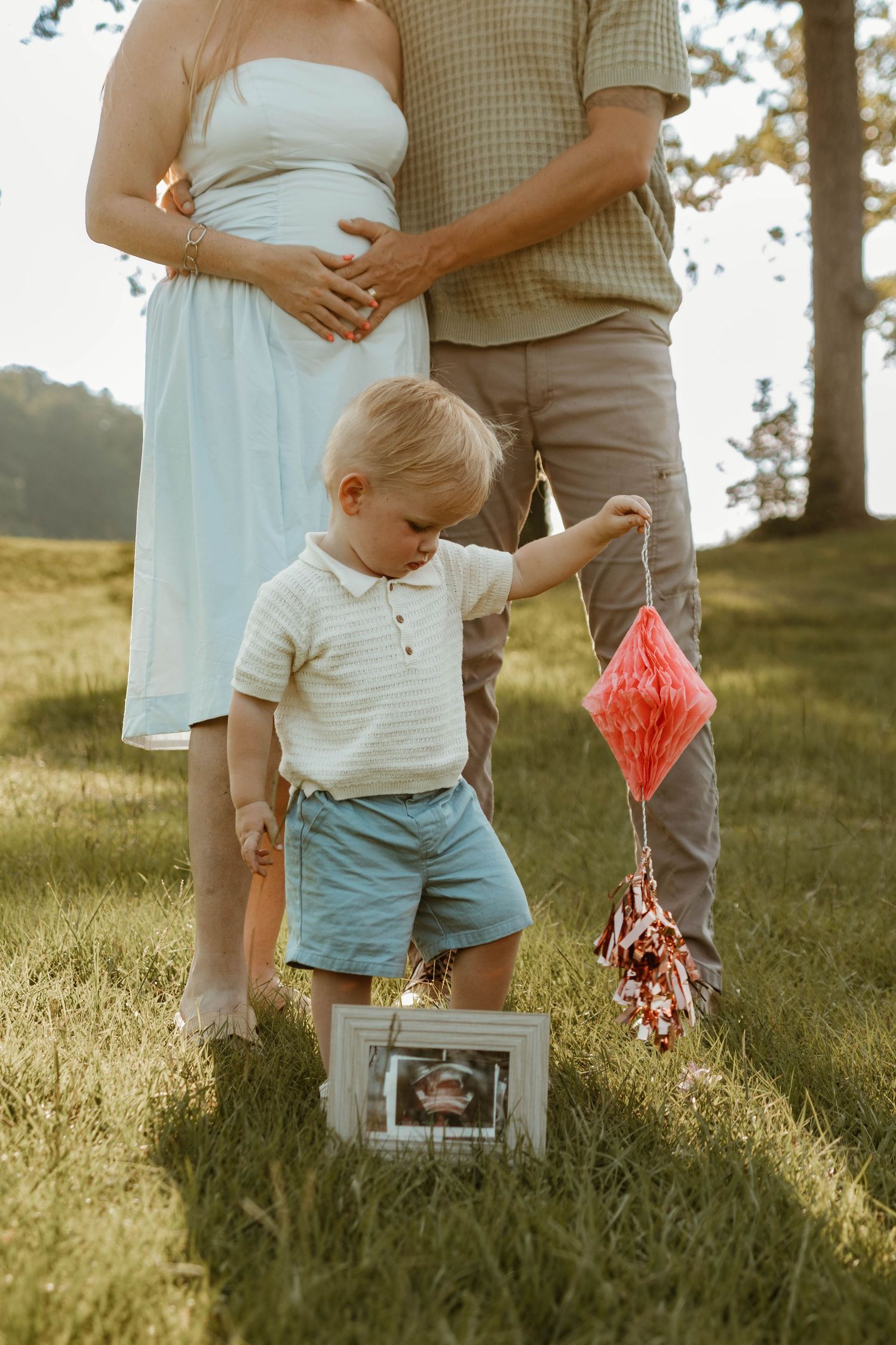 A toddler in blue shorts and white shirt holds a pink paper decoration in a sunlit grassy field in Toccoa, GA.