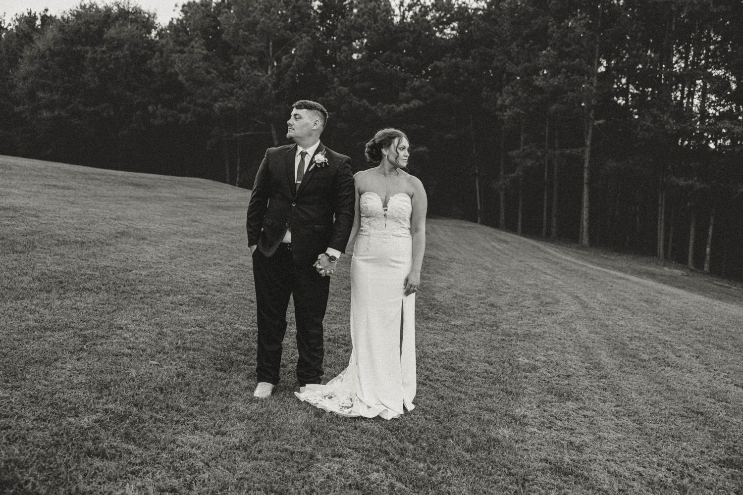 A newlywed couple stands together on a grassy hillside with trees in the background.