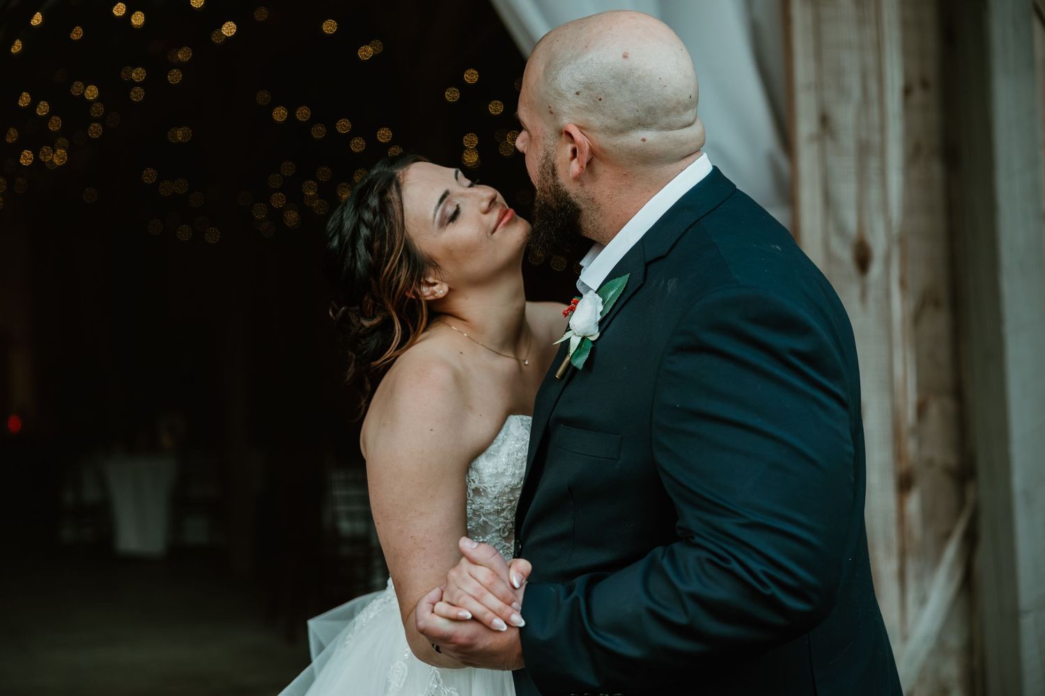 A bride and groom share a romantic kiss during their wedding reception in a rustic barn venue with warm bokeh lights.