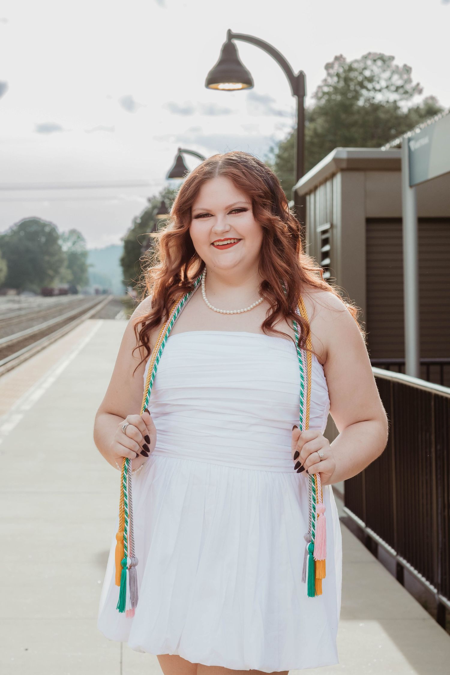 A person in a white dress with colorful tassels smiles on a train platform under a vintage lamp post in Toccoa, Georgia.