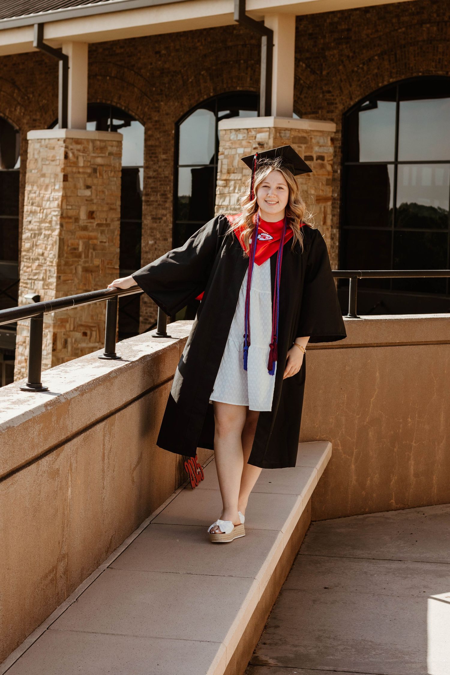 A graduate in black cap and gown with red scarf poses on a stone building ramp in Toccoa, GA.