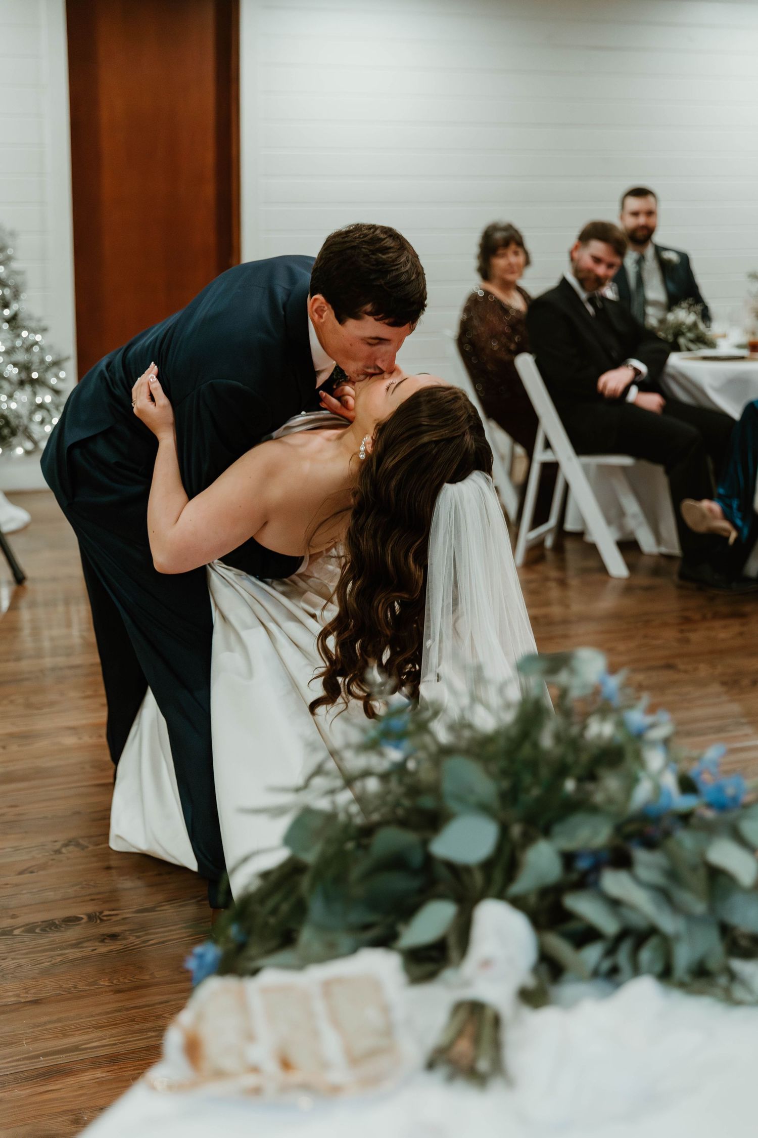 Groom dips bride for a kiss during first dance at wedding reception while guests watch in background.
