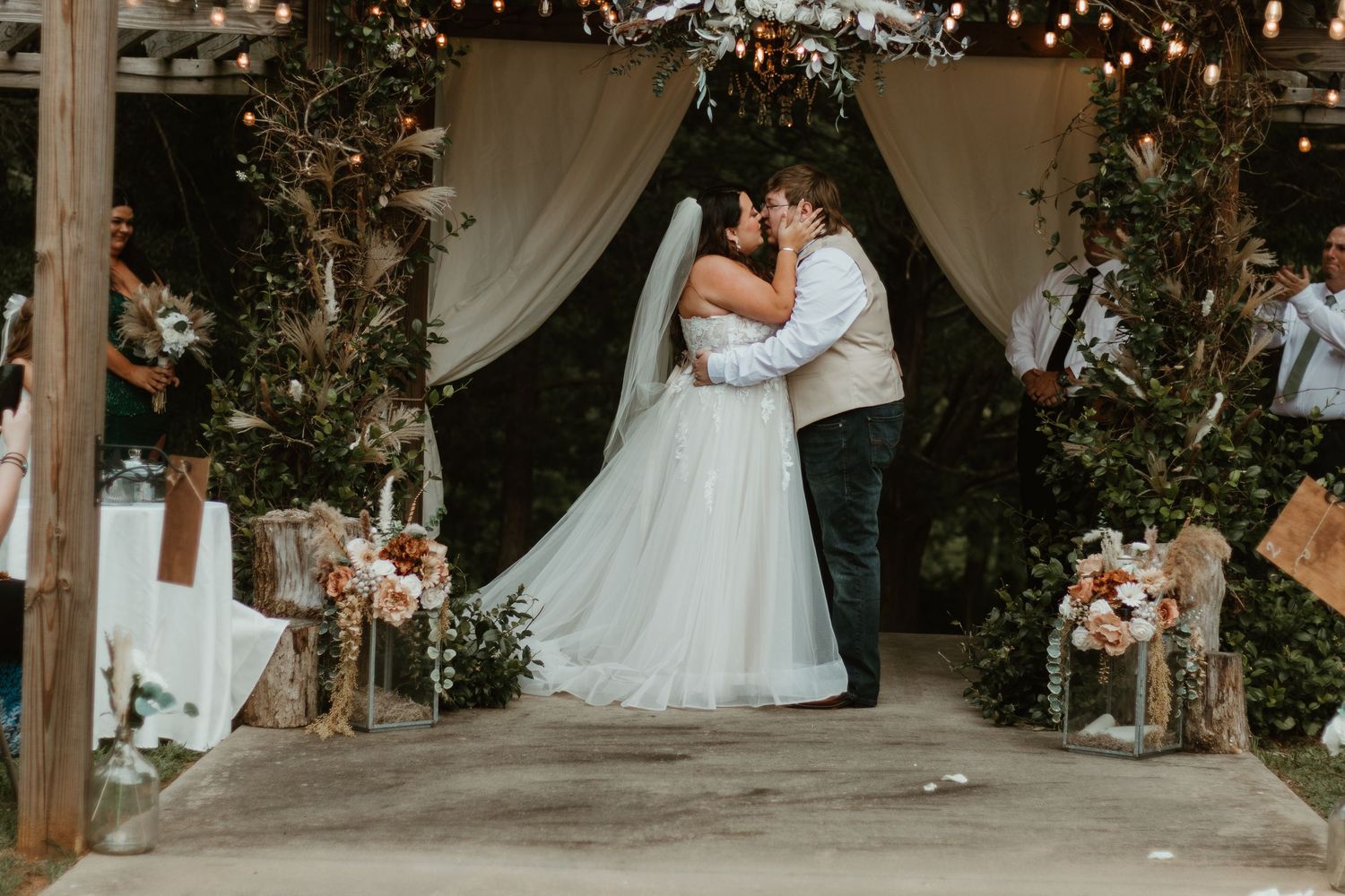 A wedding couple shares a romantic kiss under twinkling lights in an elegant rustic indoor-outdoor venue setting.