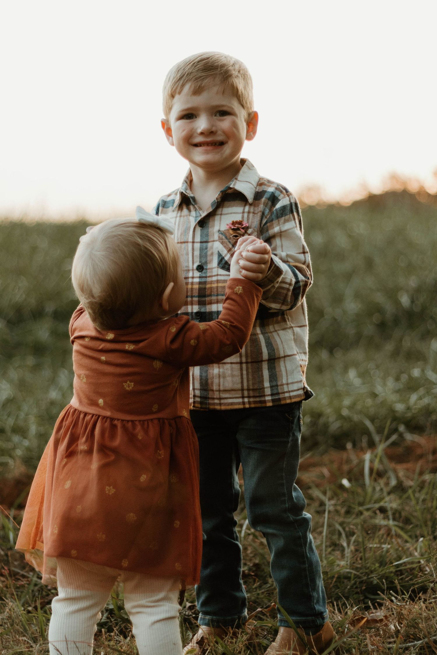 Two young children in autumn clothing interact playfully in a grassy field at sunset.