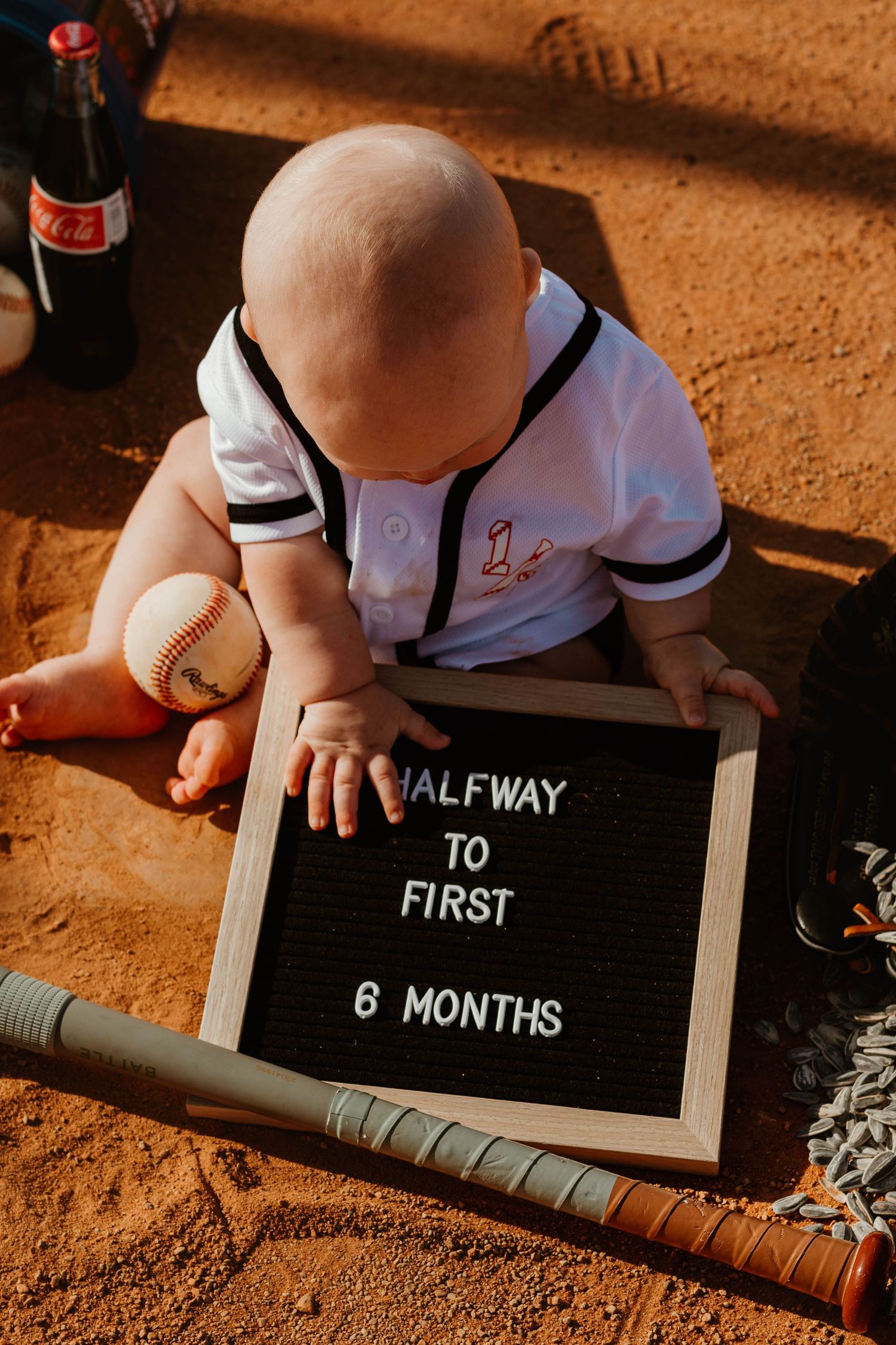 Baby in baseball outfit sits with milestone sign reading 'Halfway to First 6 Months' on dirt ground in northeast Georgia.