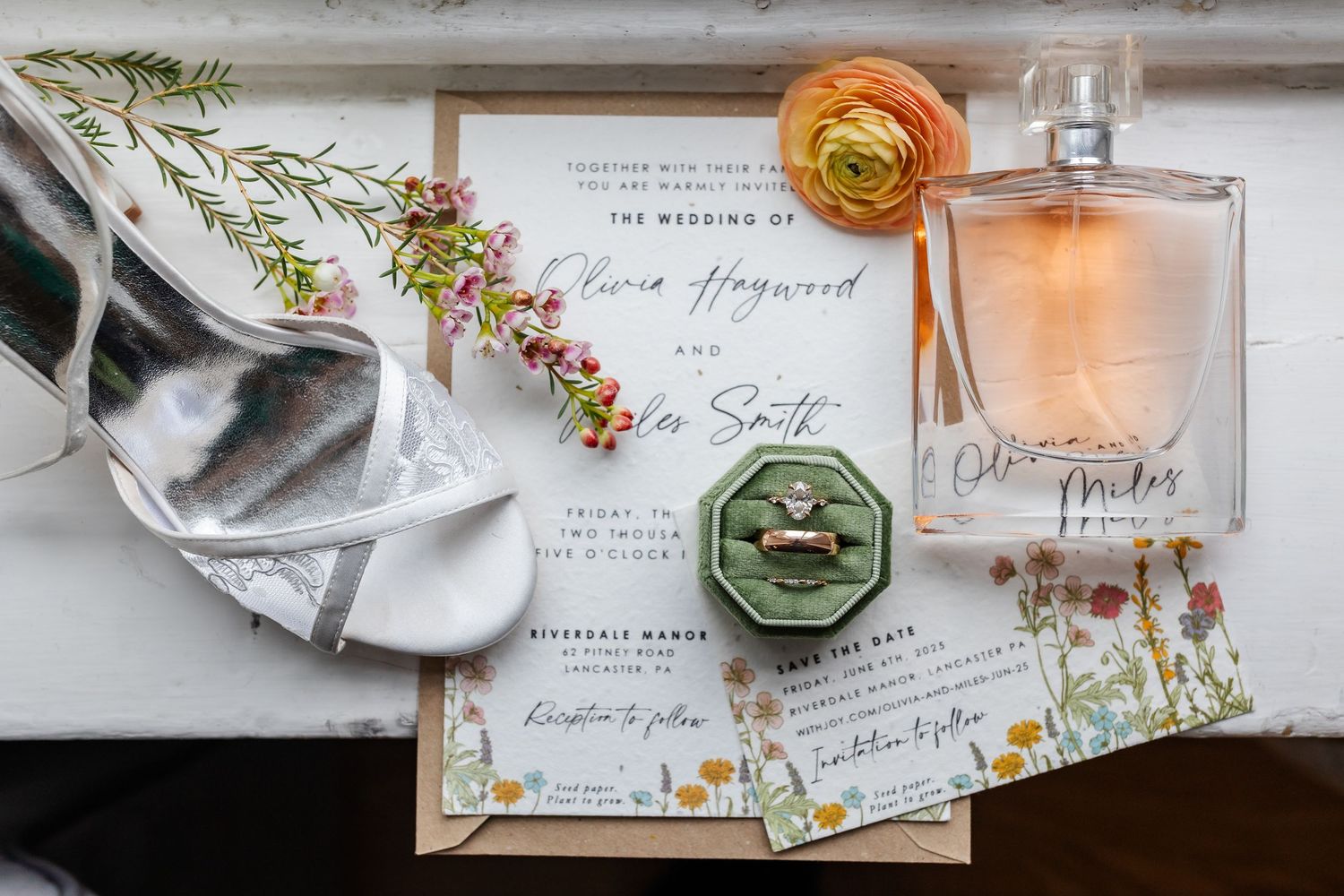 Wedding invitation details with perfume bottle, orange ranunculus flower, and sage green wax seal on white fabric.