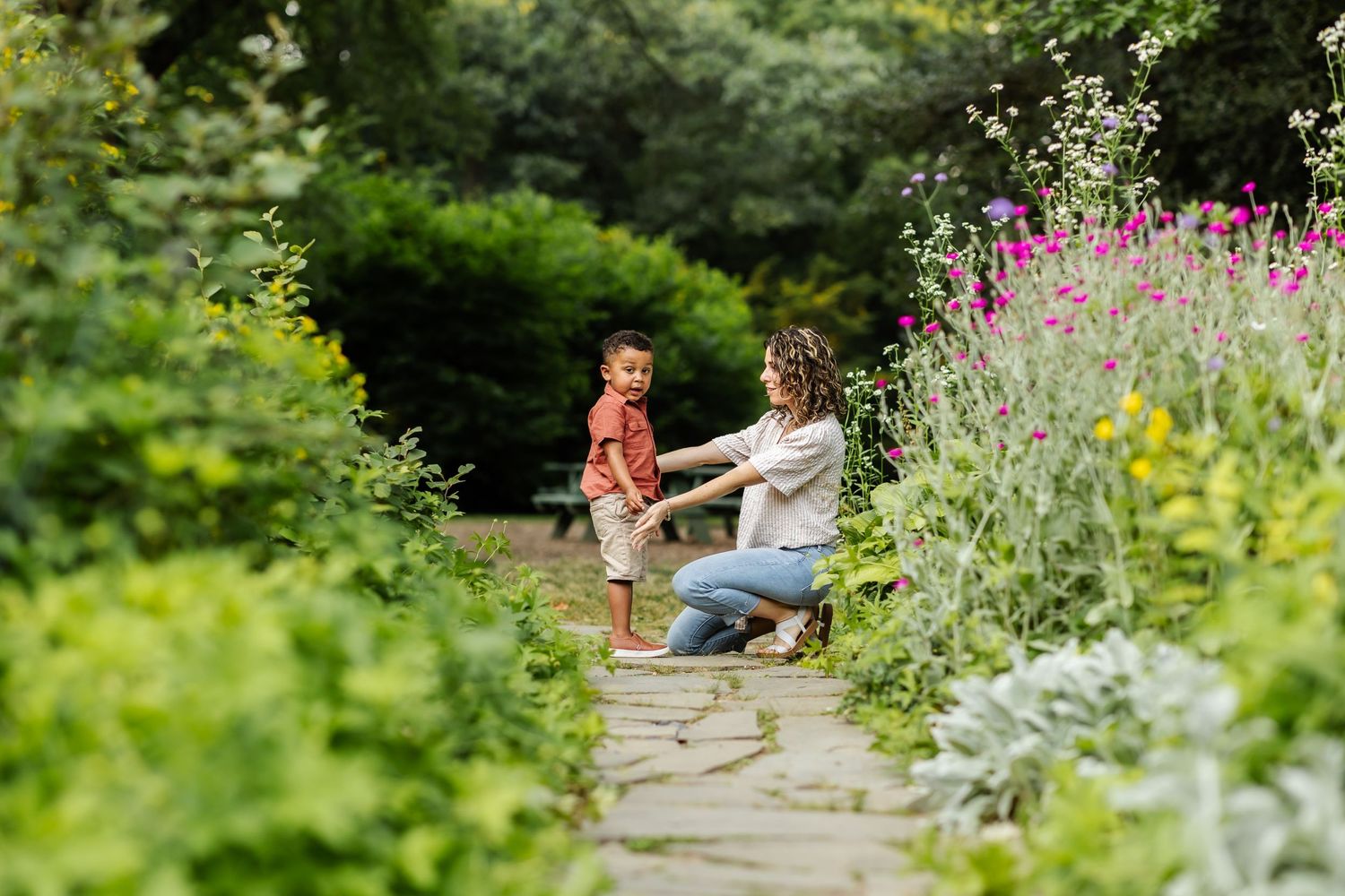 A mom reaches out for a hug from her toddler son in a flowering garden