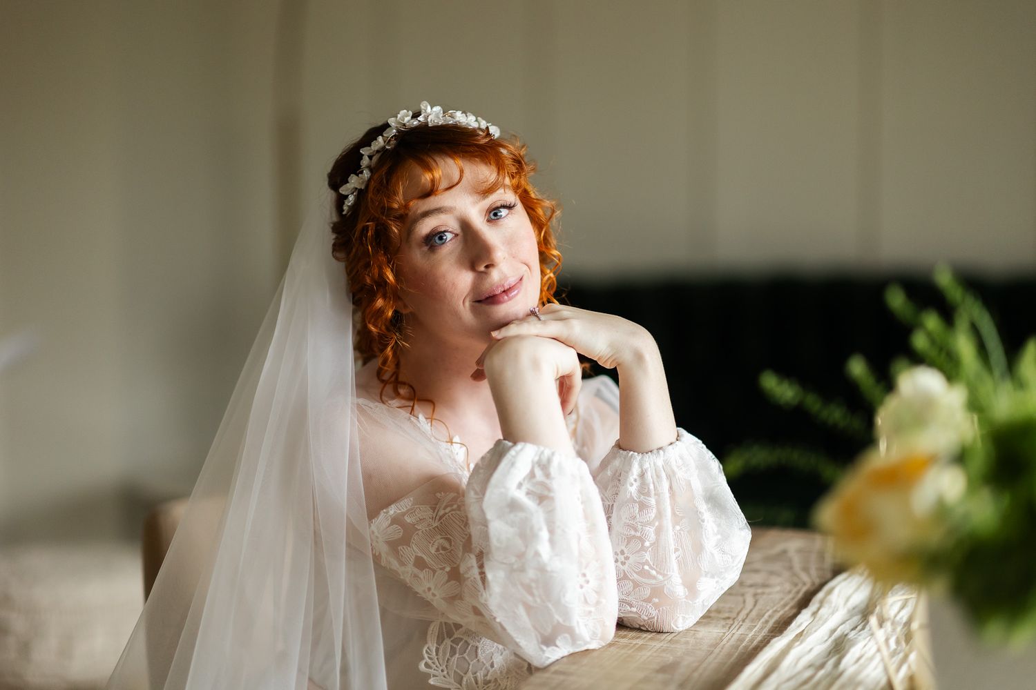A bride in a vintage-style lace wedding dress with long sleeves and veil rests her chin thoughtfully on her hand.