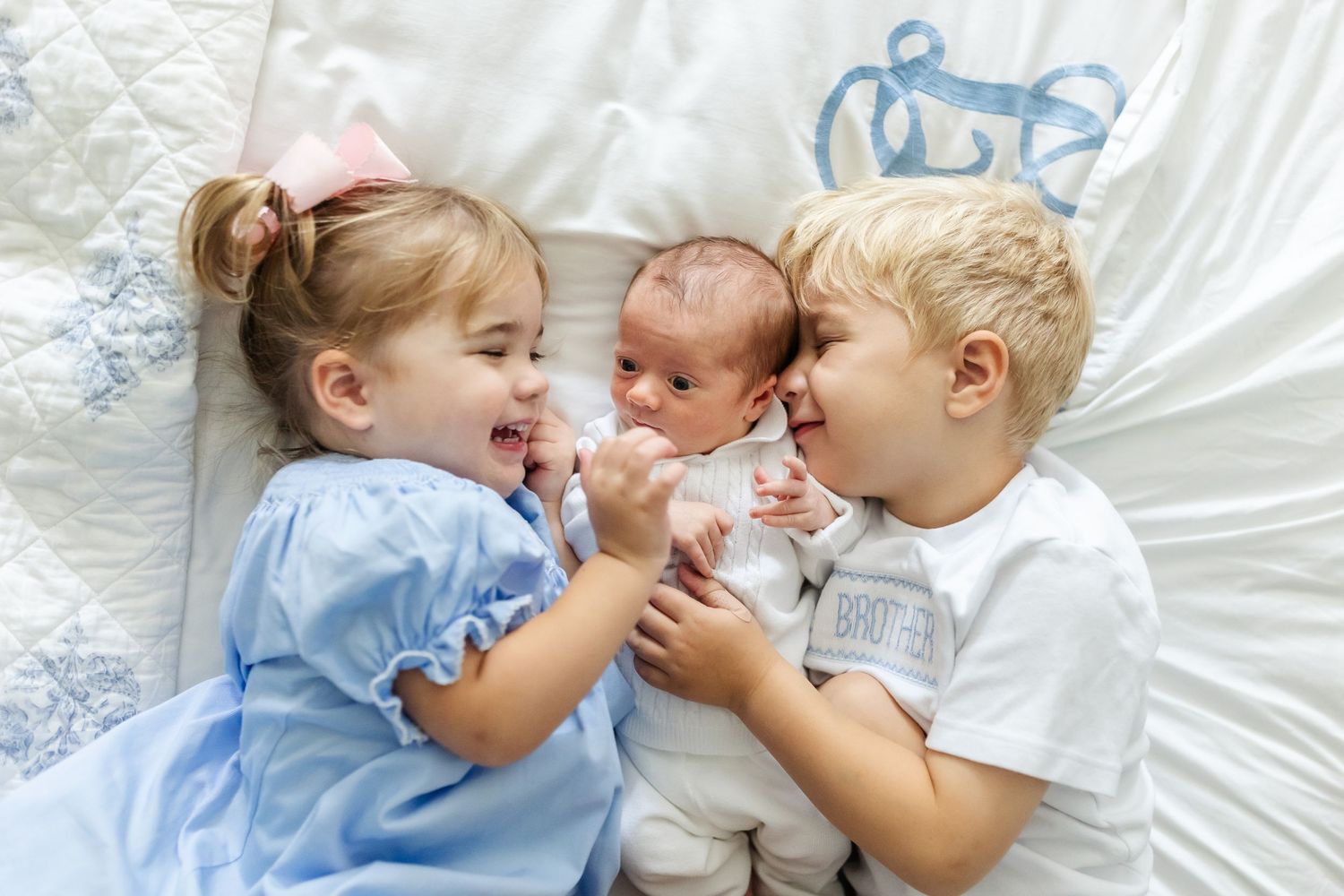 Three young children lying on white bedding together, smiling and playing.