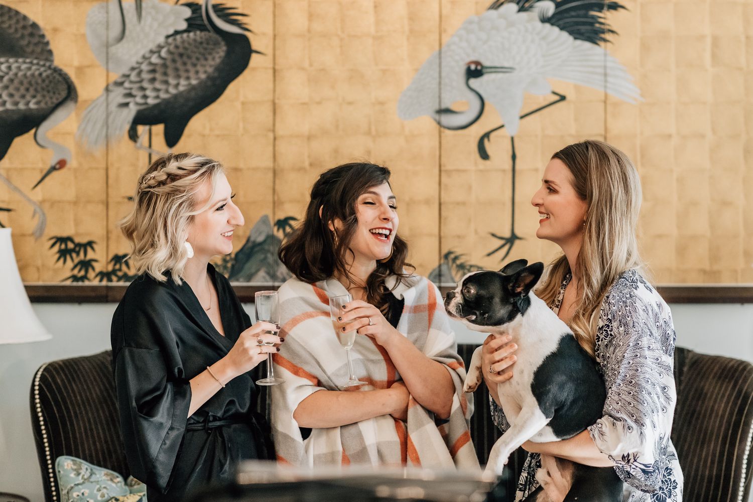 Three friends laughing together on a sofa while holding small dogs with Asian-inspired artwork on the wall behind them.