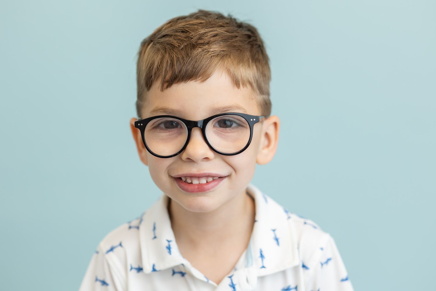 Smiling young boy wearing large black glasses and a white patterned shirt against a light blue background.
