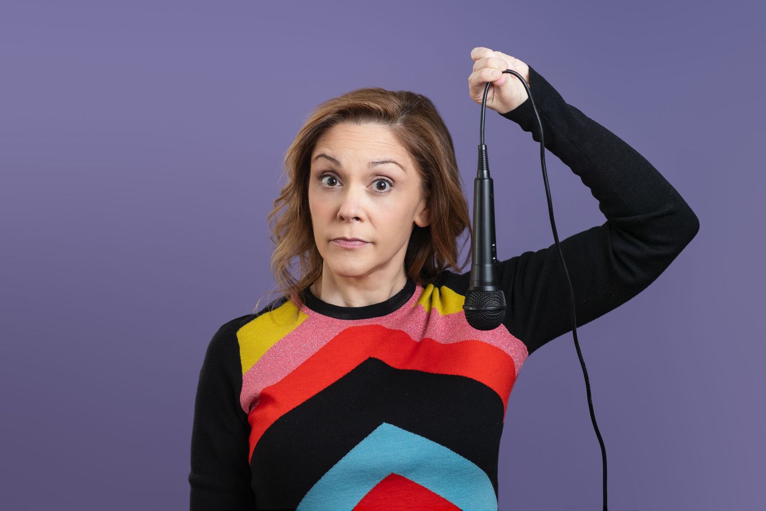 Someone wearing a colorful geometric patterned sweater holds up a black cord against a purple background.