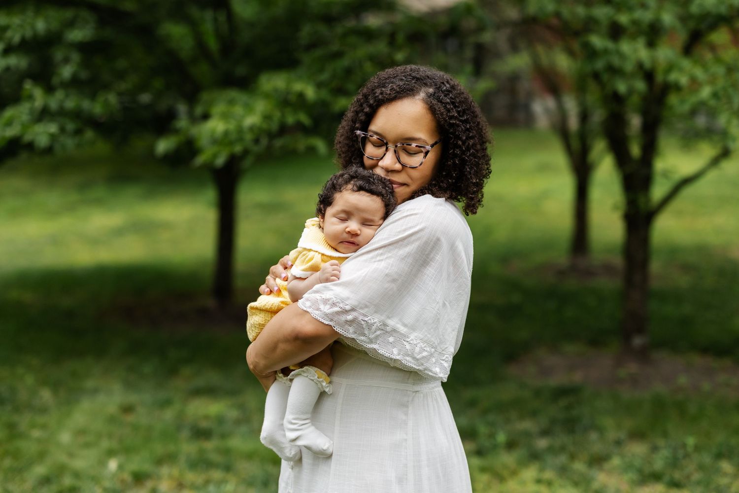 Parent in white dress holds sleeping infant outdoors in a lush green park setting.