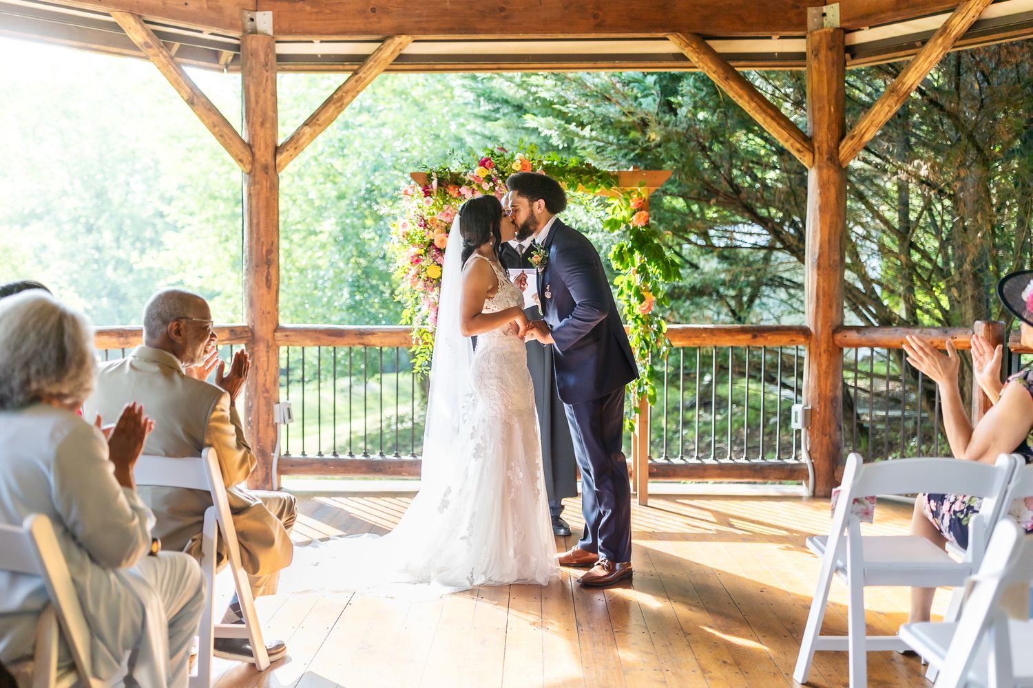 A wedding ceremony takes place under a rustic wooden pavilion surrounded by trees and natural light.