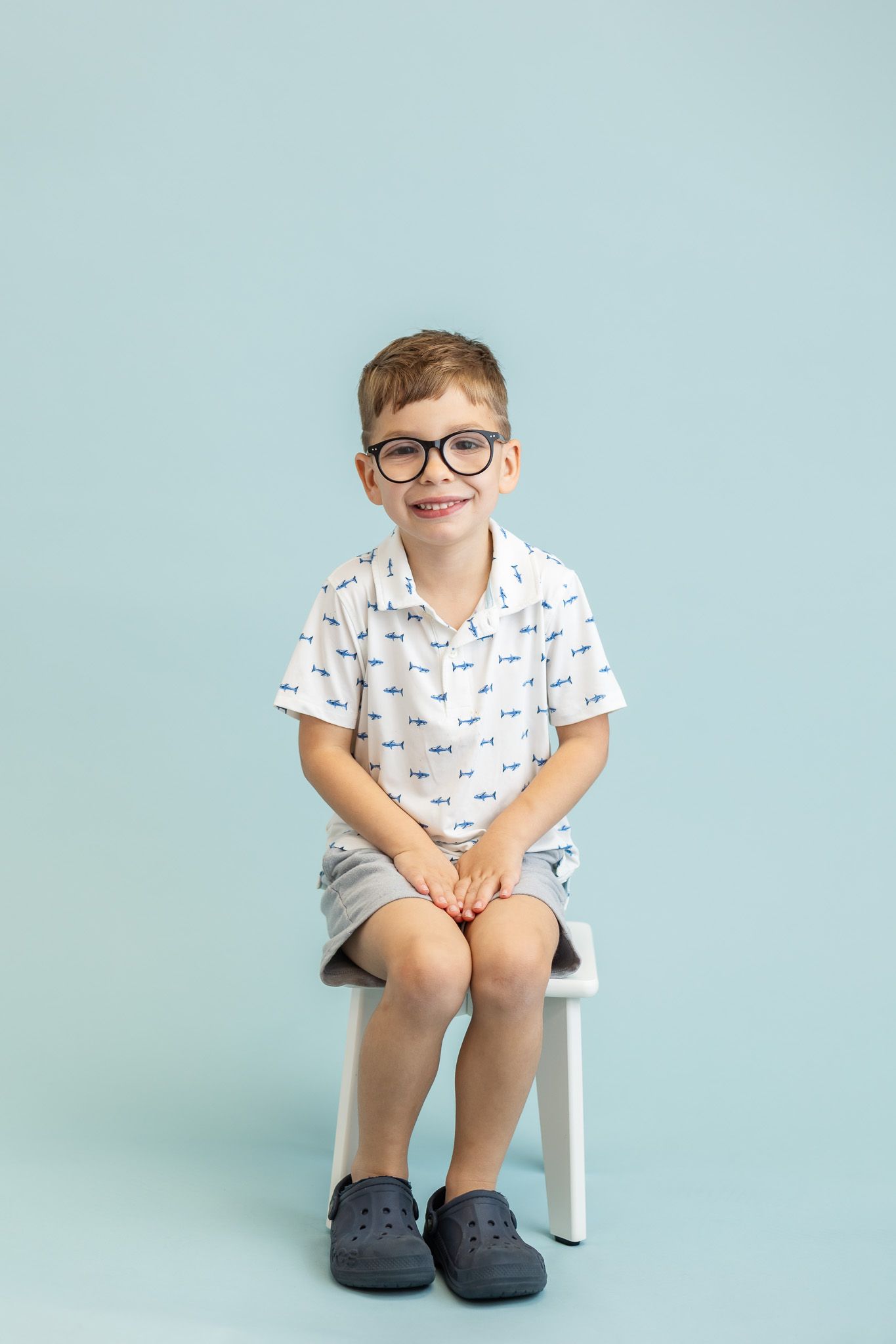 Smiling young boy wearing glasses and a patterned shirt, sitting on a white chair against a light blue background.