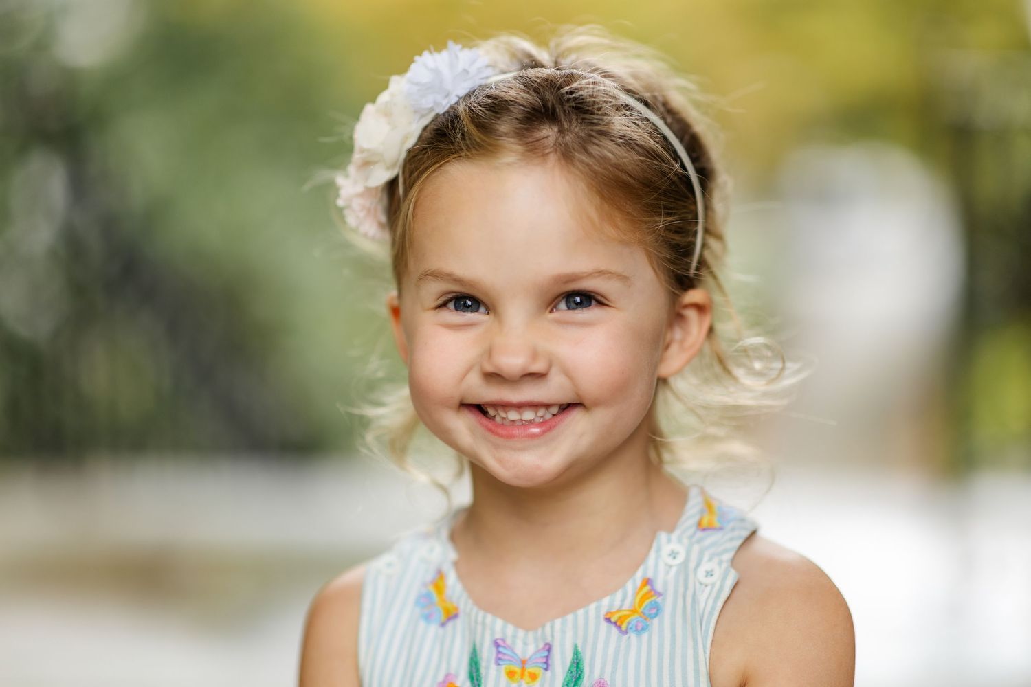 Smiling young blonde girl wearing a floral dress and white headband, posing outdoors with a blurred green background.