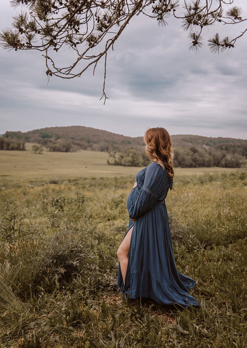 Pregnant woman stands in a field wearing a long blue dress