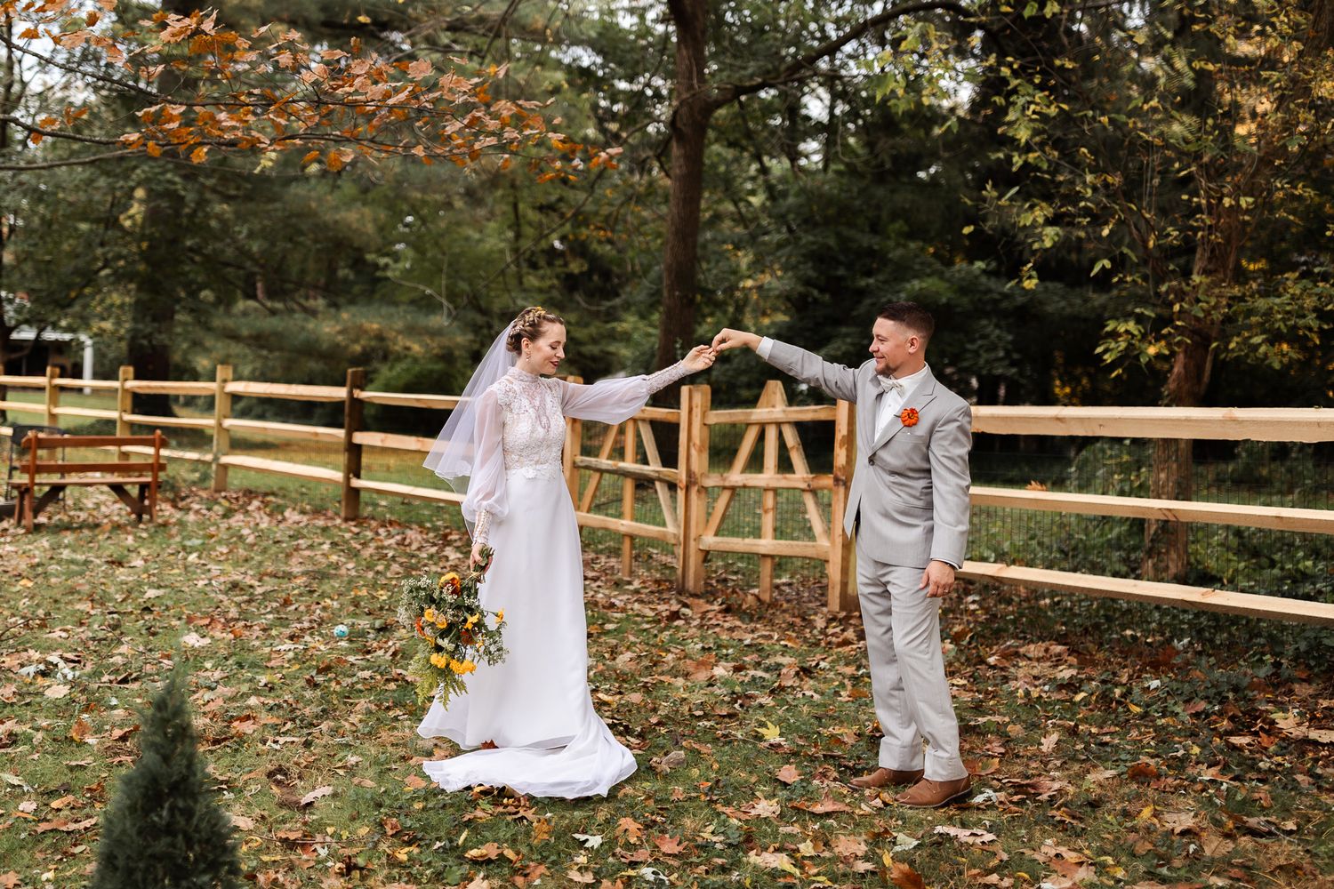 A couple in wedding attire shares playful moments in a fall setting with wooden fences and autumn leaves on the ground.