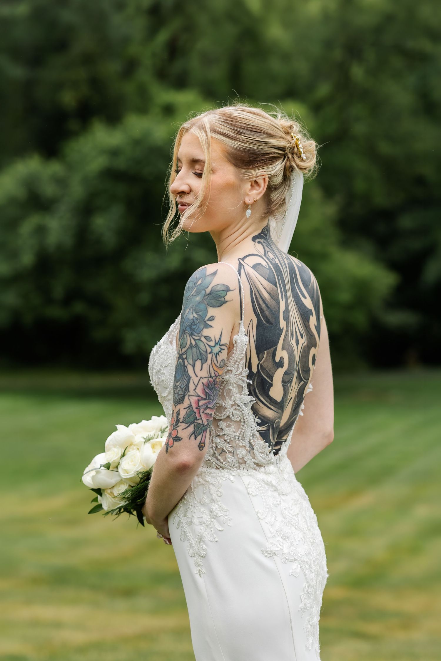 Tattooed bride in backless lace wedding gown holding white peony bouquet outdoors in garden setting.