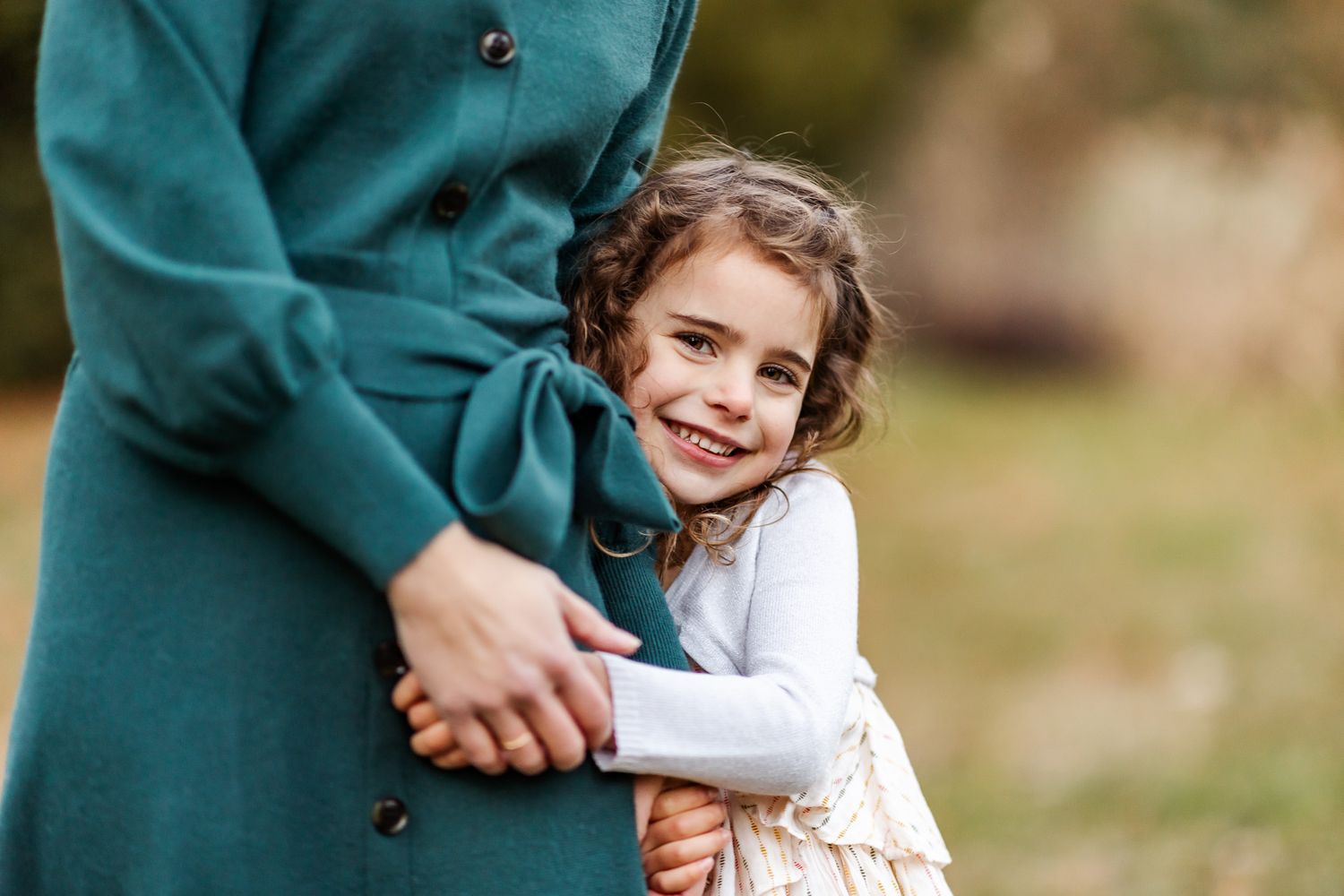 Child wearing white dress embraced in teal coat during outdoor portrait session.