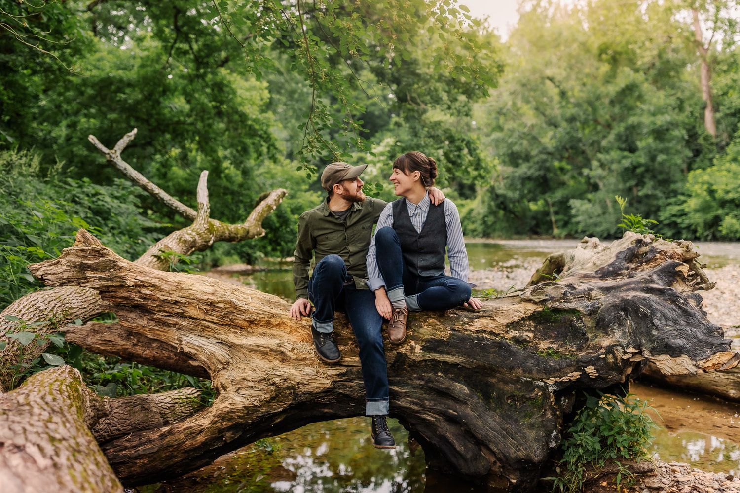 Young couple sits on a fallen tree by the creek while smiling at each other