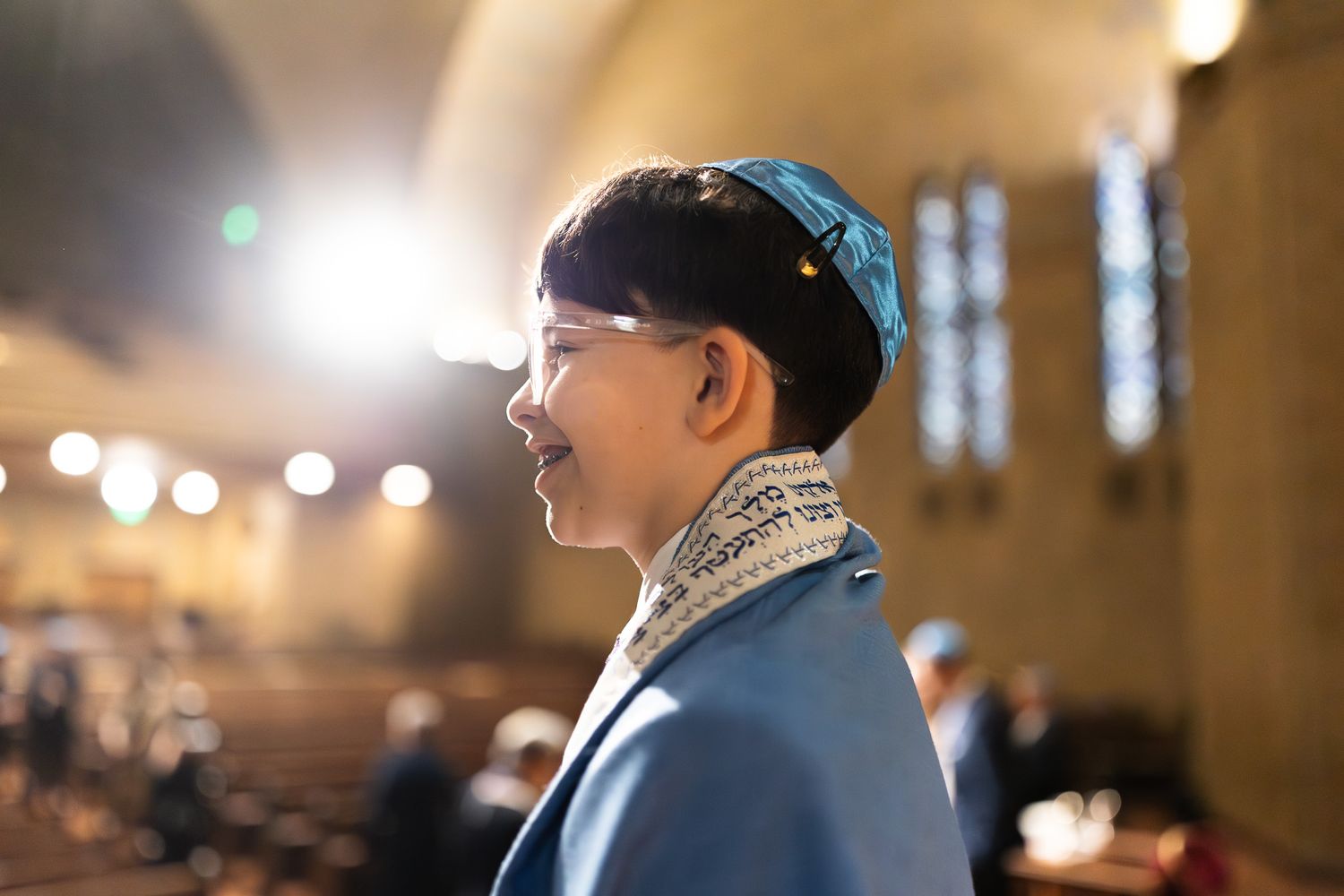 Young person wearing a blue kippah smiling in profile inside an ornate synagogue during a Bar Mitzvah.