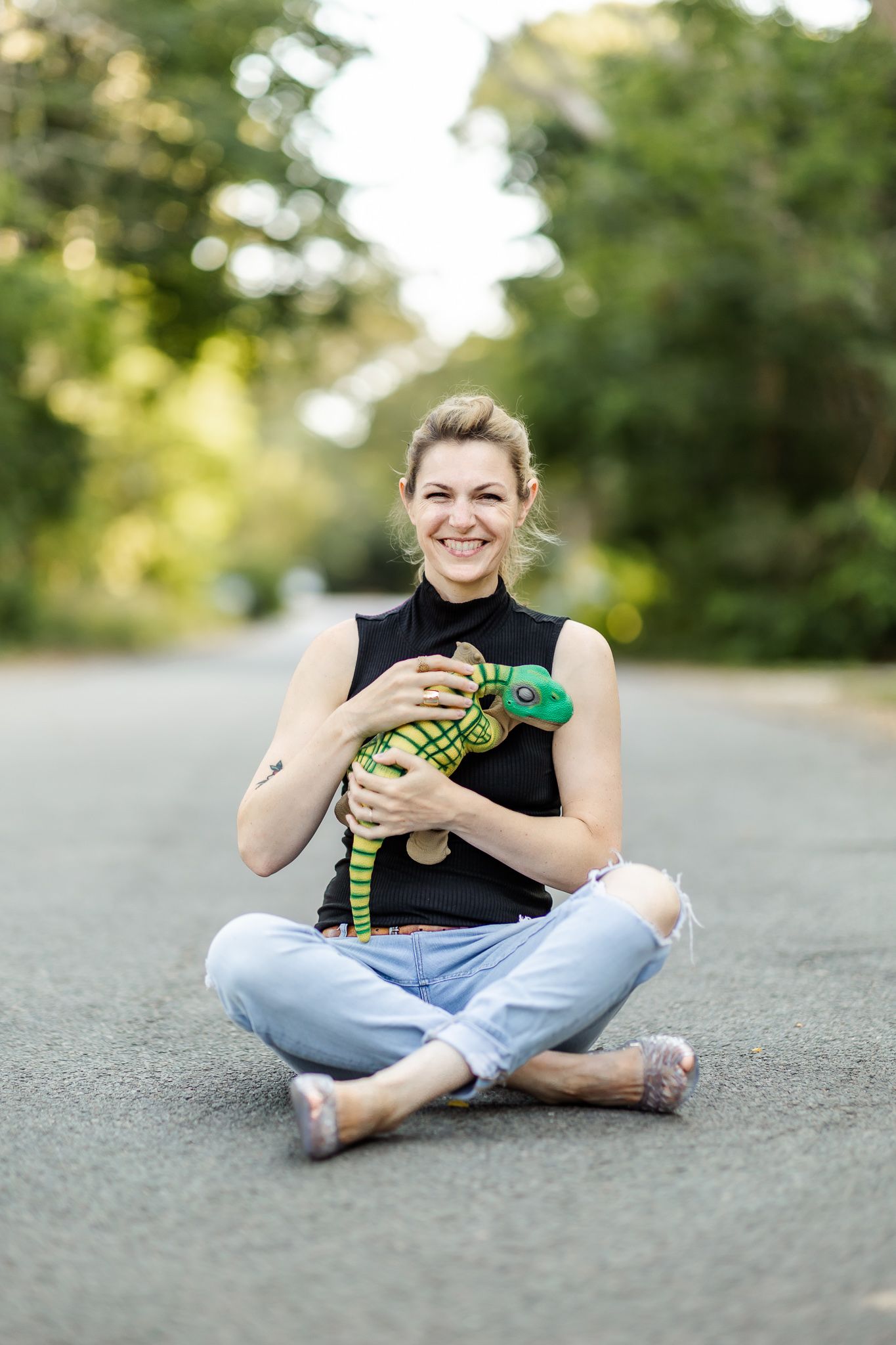 Researcher Kate Darling smiles broadly and sits in the road with her small dinosaur robot