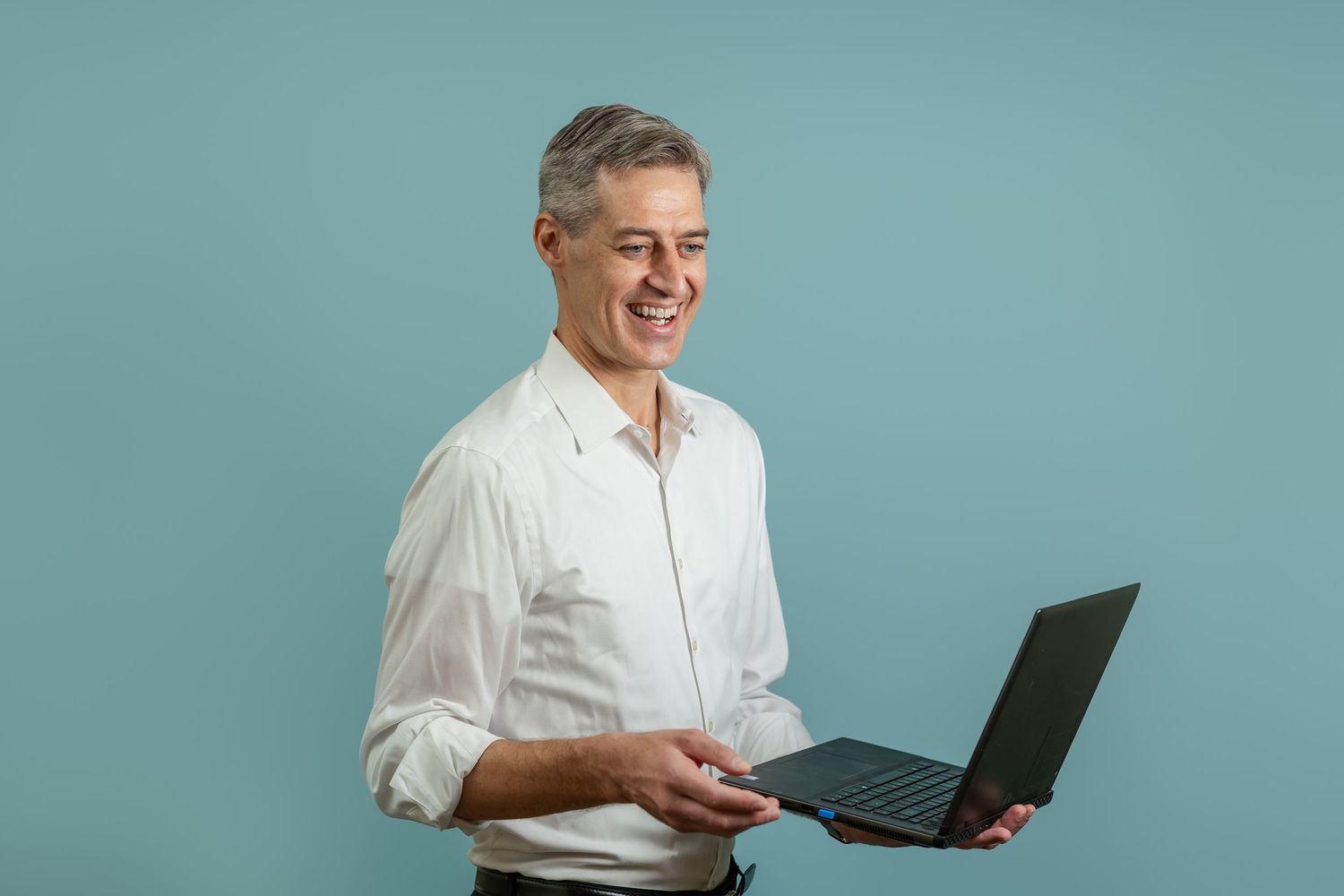 Professional in white shirt smiles while holding and working on a laptop against turquoise background.