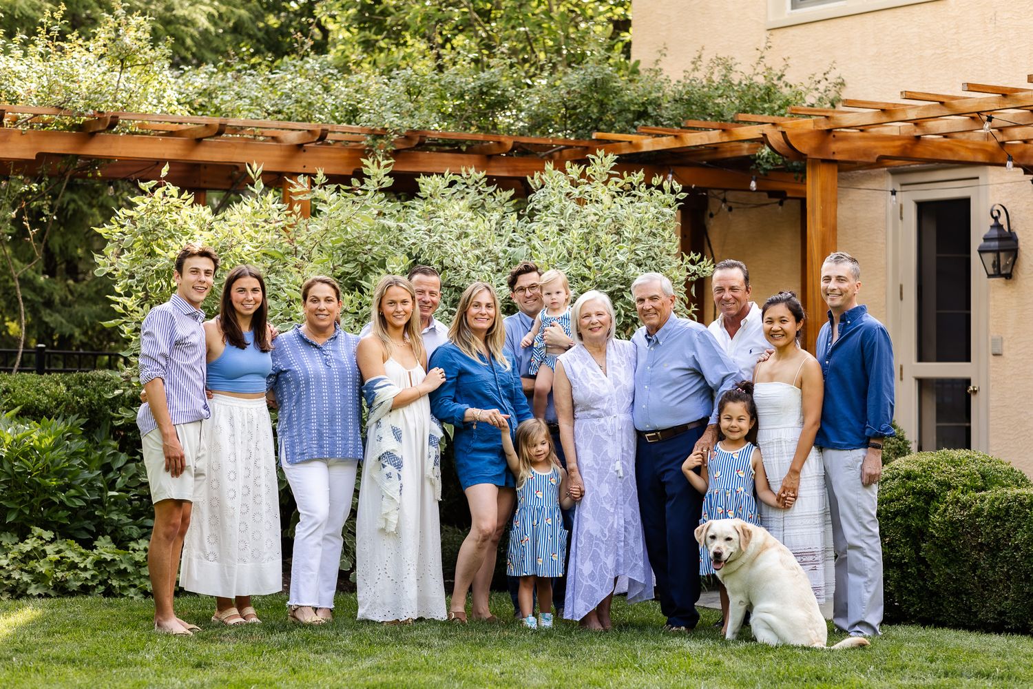 Large multigenerational family group portrait outdoors with pergola, lush greenery, and a golden retriever dog on sunny day.