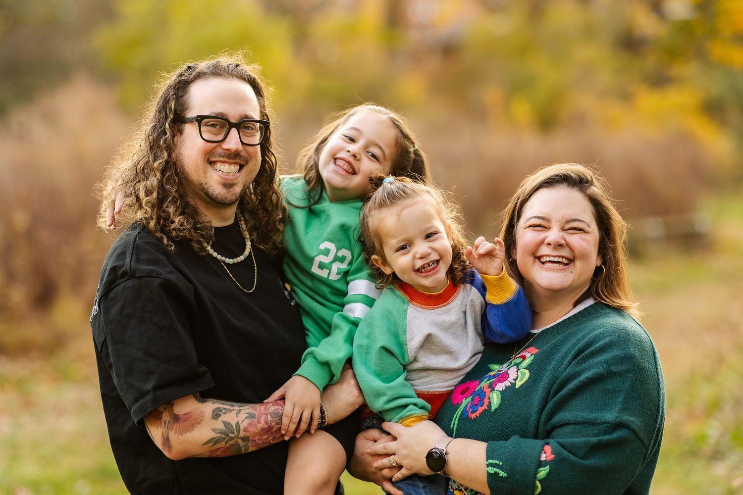 Happy family of four laughing outdoors in autumn, parents holding two young children in a colorful fall setting.