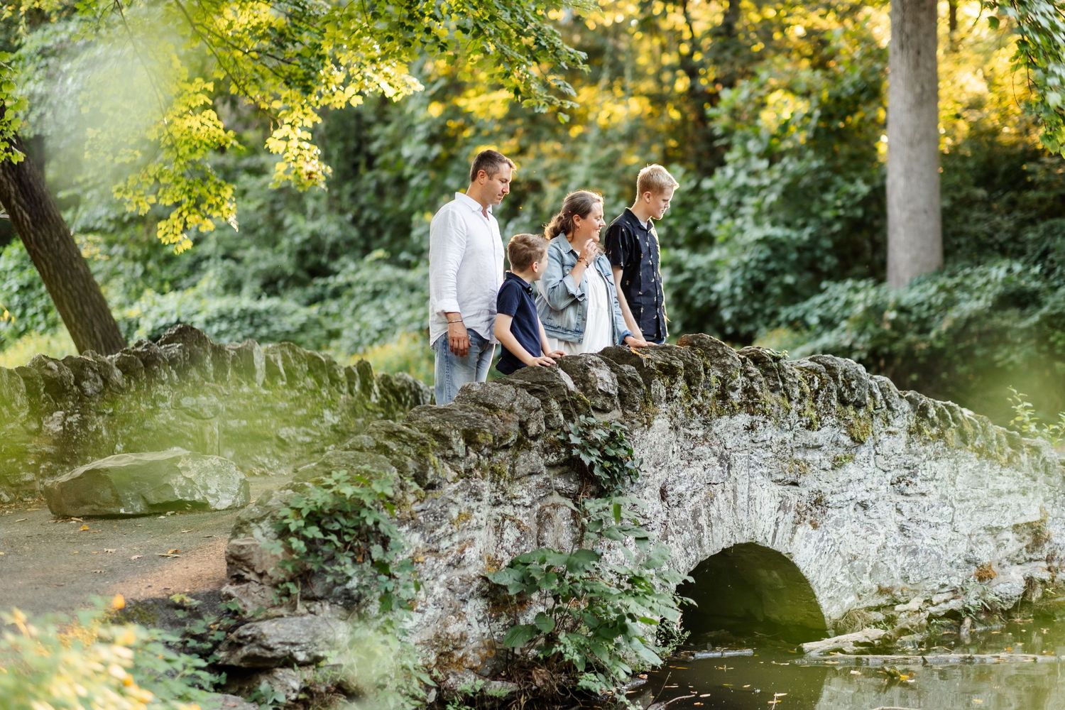 A family with two sons stands on a stone bridge over a calm stream