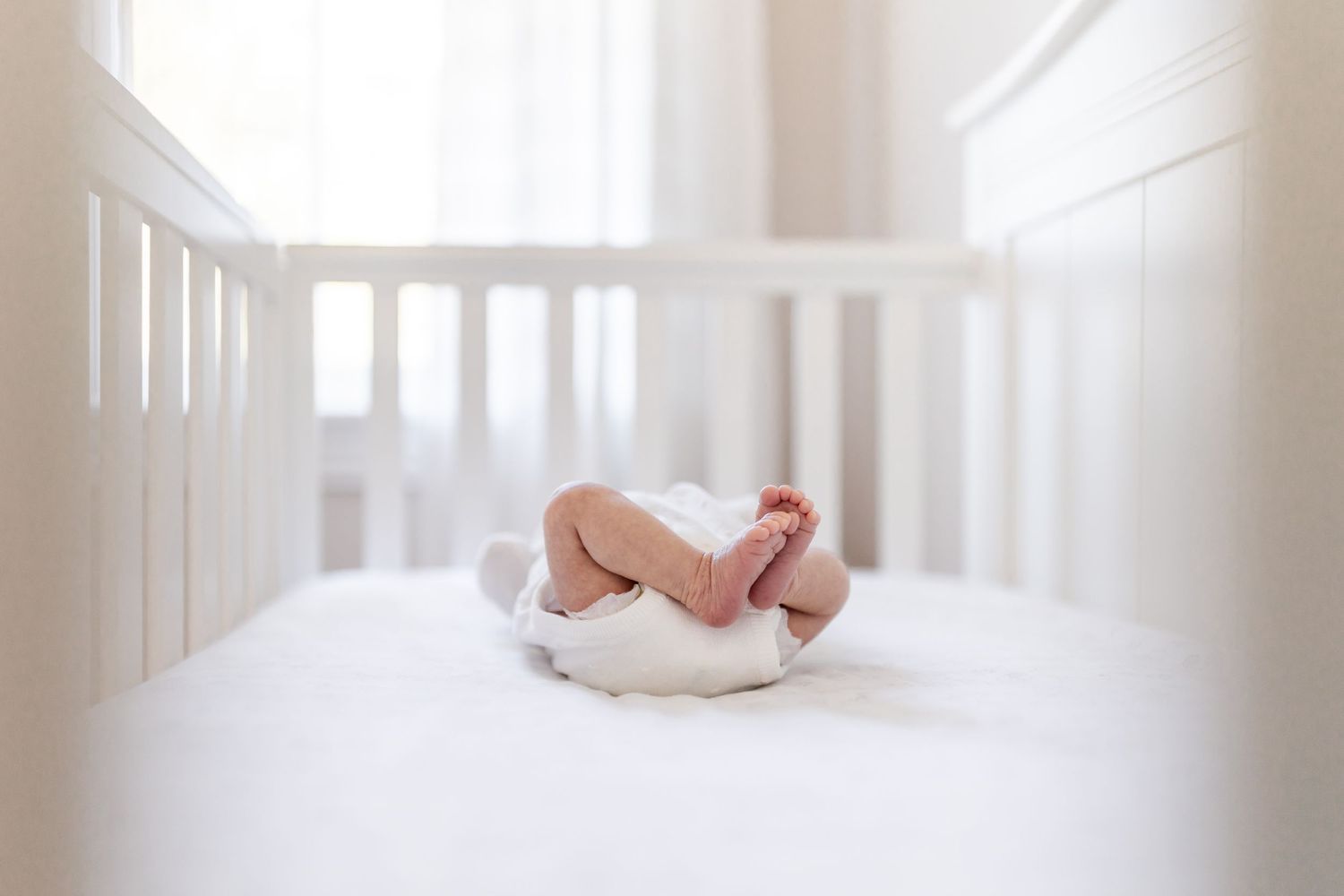 Baby resting on white bedding inside a white crib with natural light.