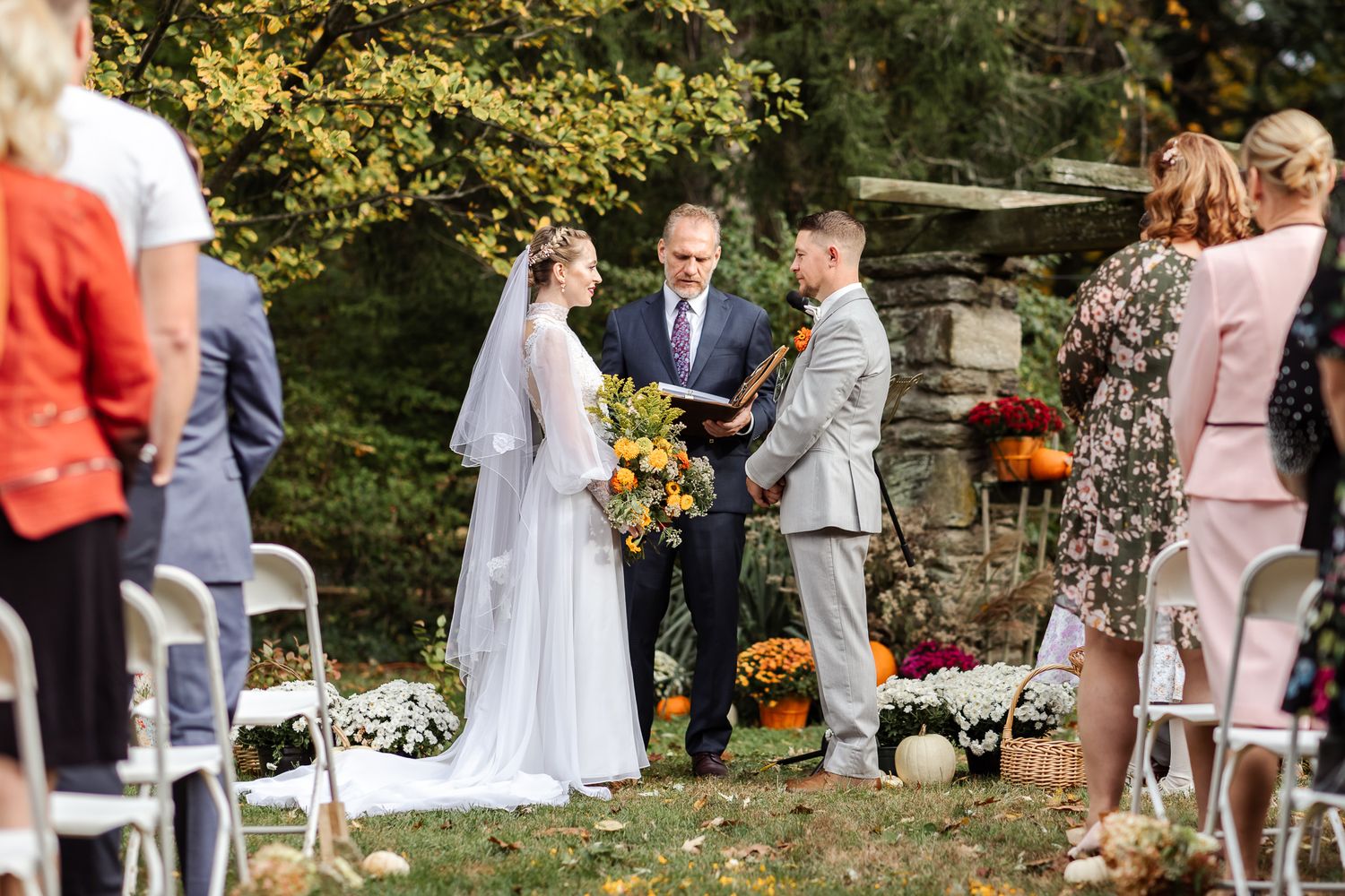 Wedding guests gather in a garden setting with colorful autumn flowers and stone walls during an outdoor ceremony.