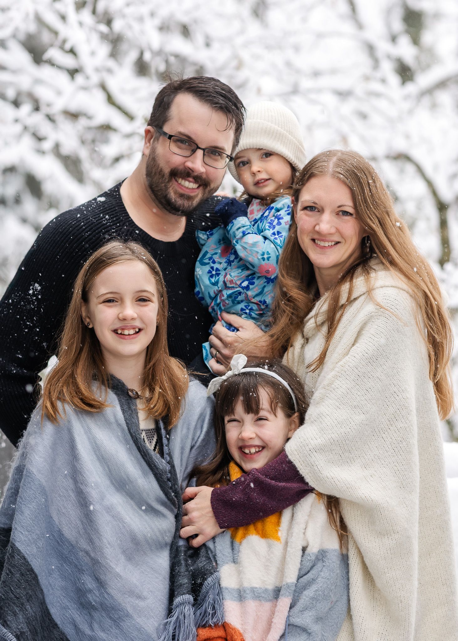 A family poses together outdoors in winter with snow falling around them against a snowy background.
