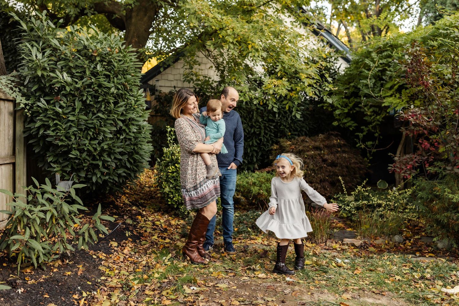 A family of four smiles as their young daughter spins around in their autumnal backyard