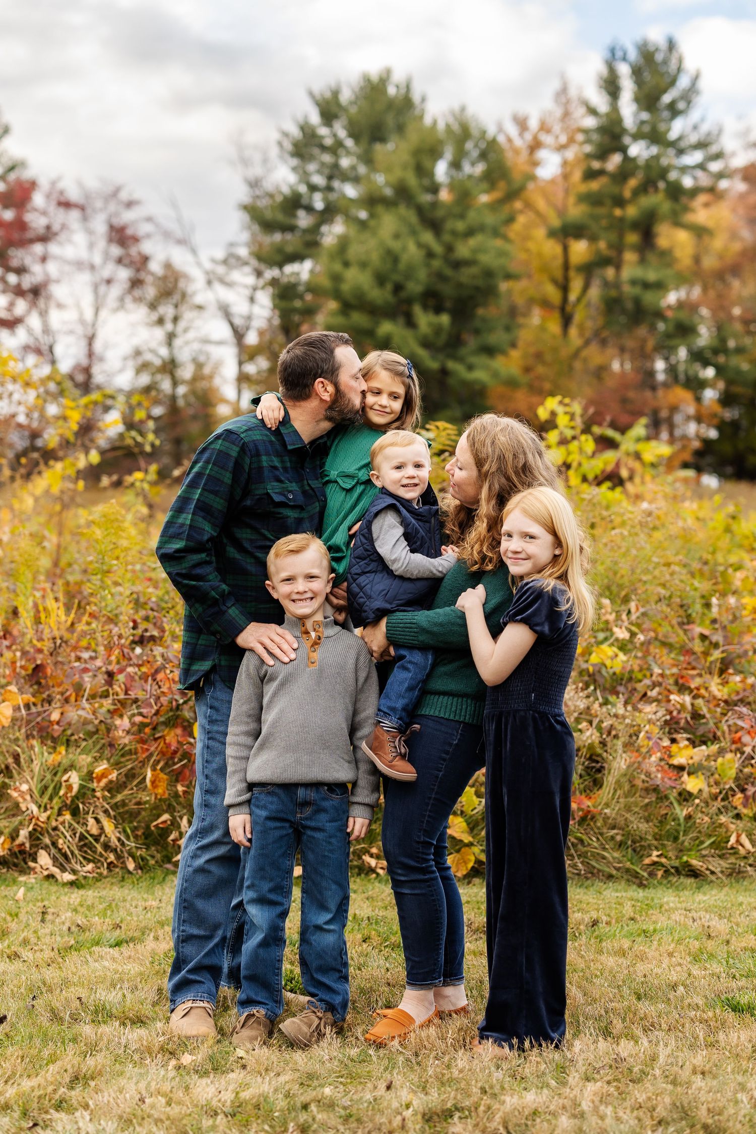Family enjoying playful moments together in an autumn field with golden foliage and evergreen trees in background.