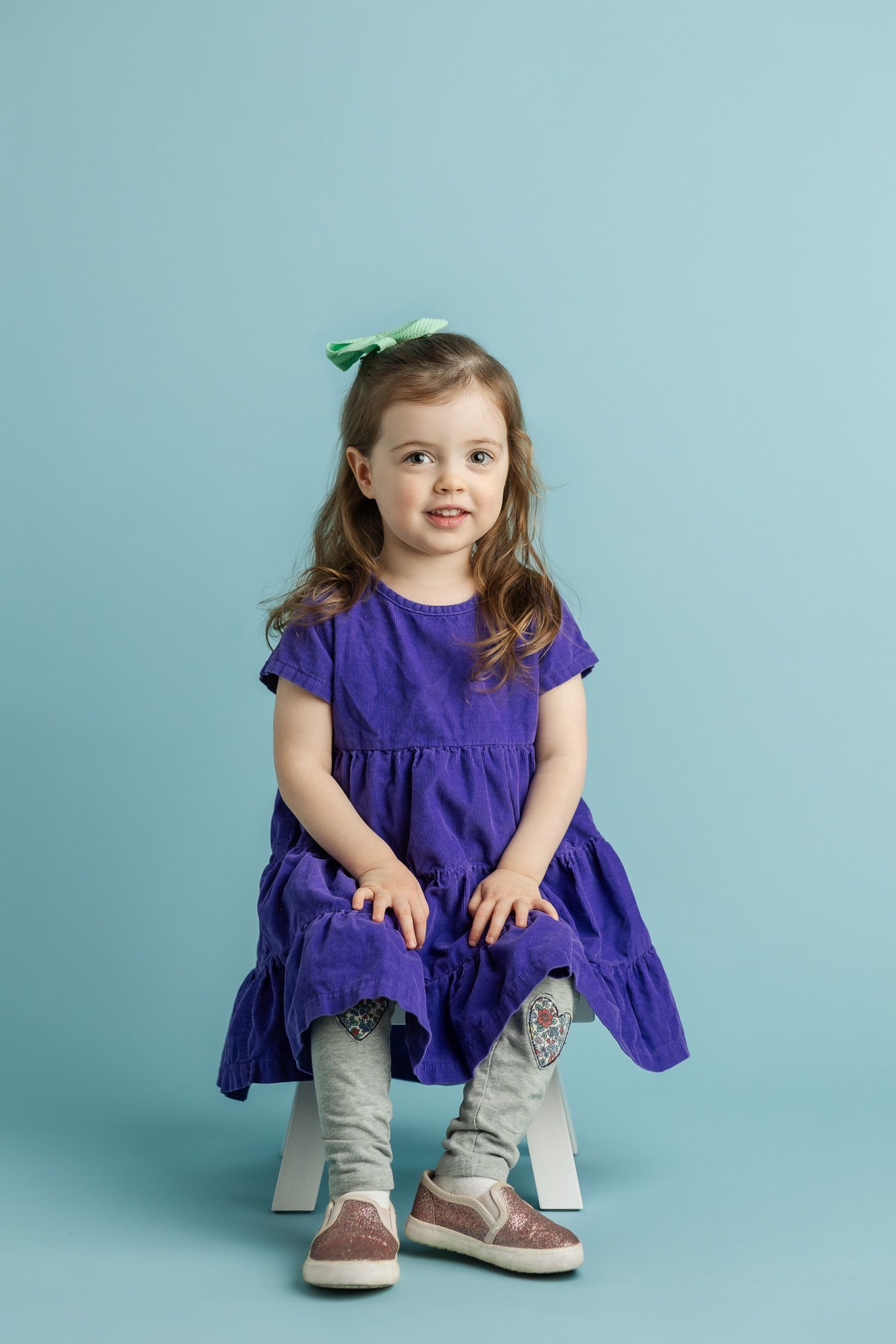 Cute toddler girl in purple dress and gray leggings with green bow sits on white stool against blue background.