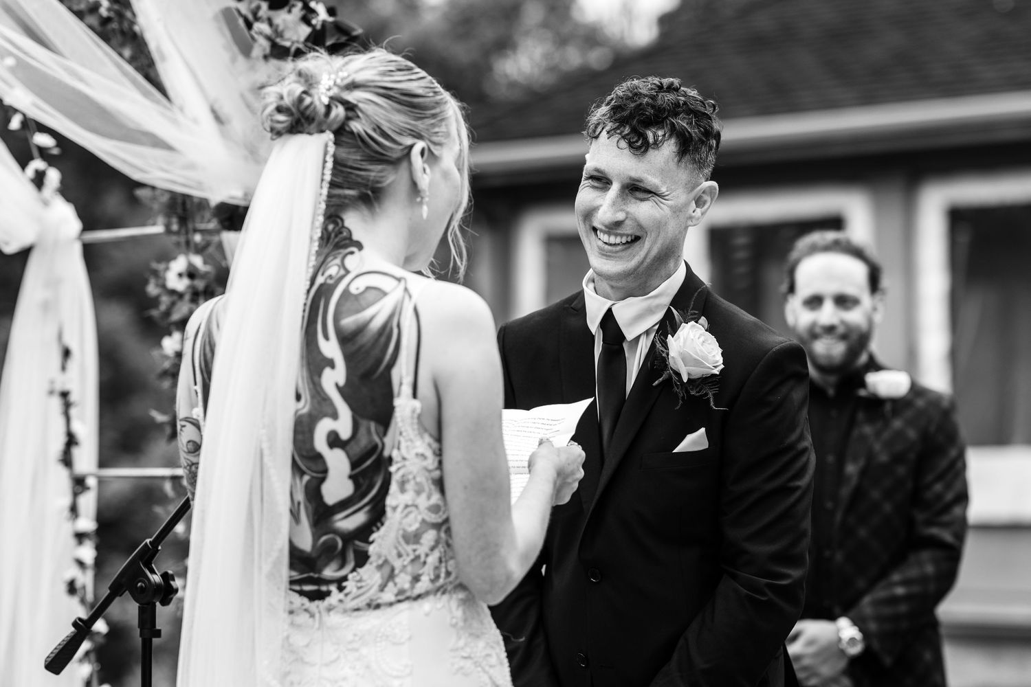 Groom smiling joyfully at bride during outdoor wedding ceremony in black and white photo.