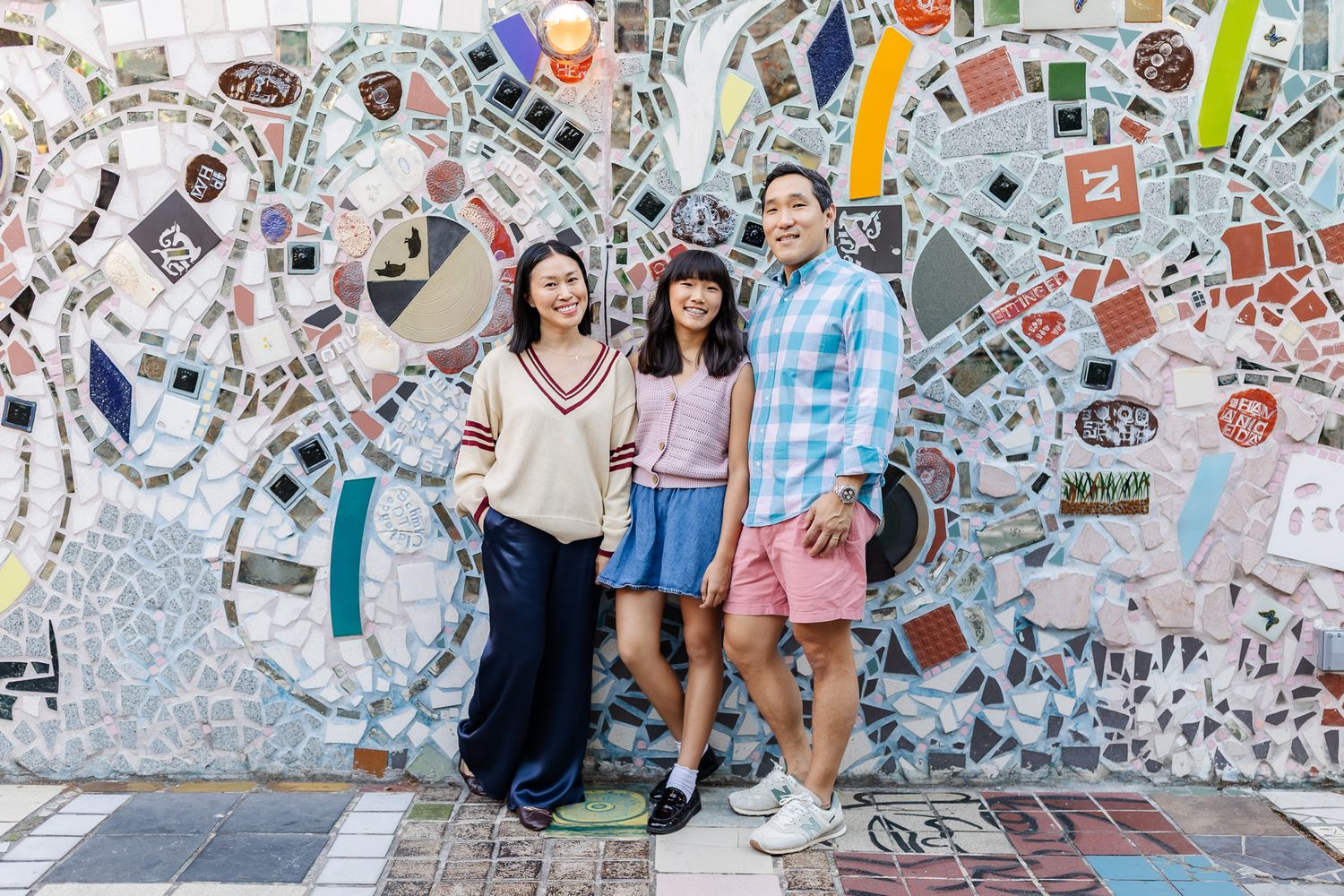 Three people smiling in front of a colorful mosaic tile wall outdoors.
