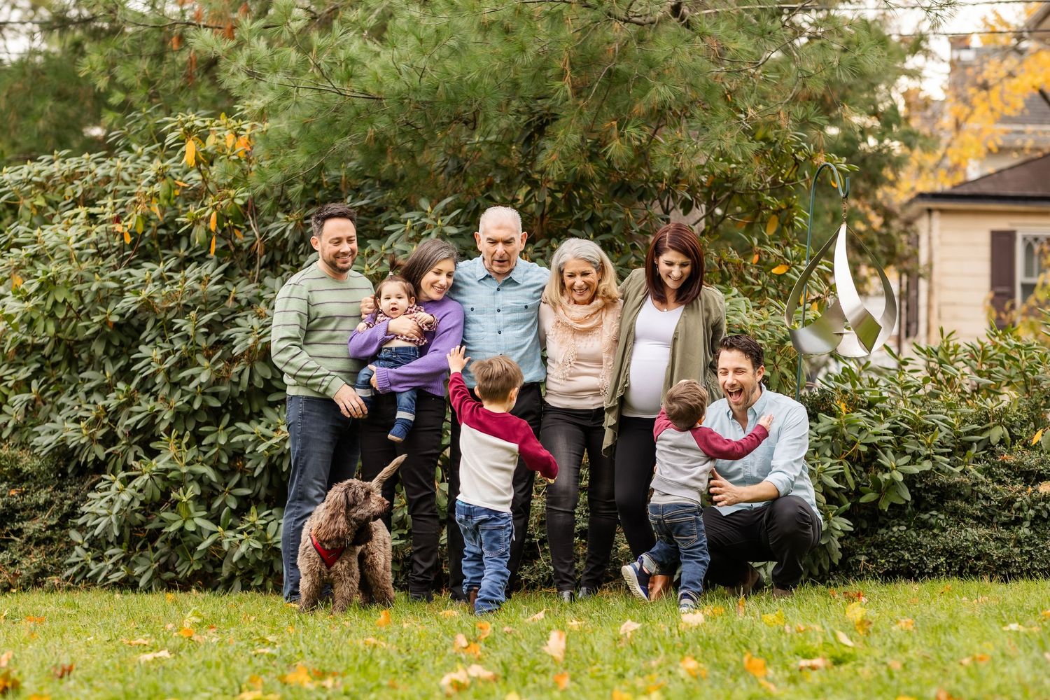Multi-generational family with dog smiling outdoors in autumn garden with lush green shrubs.