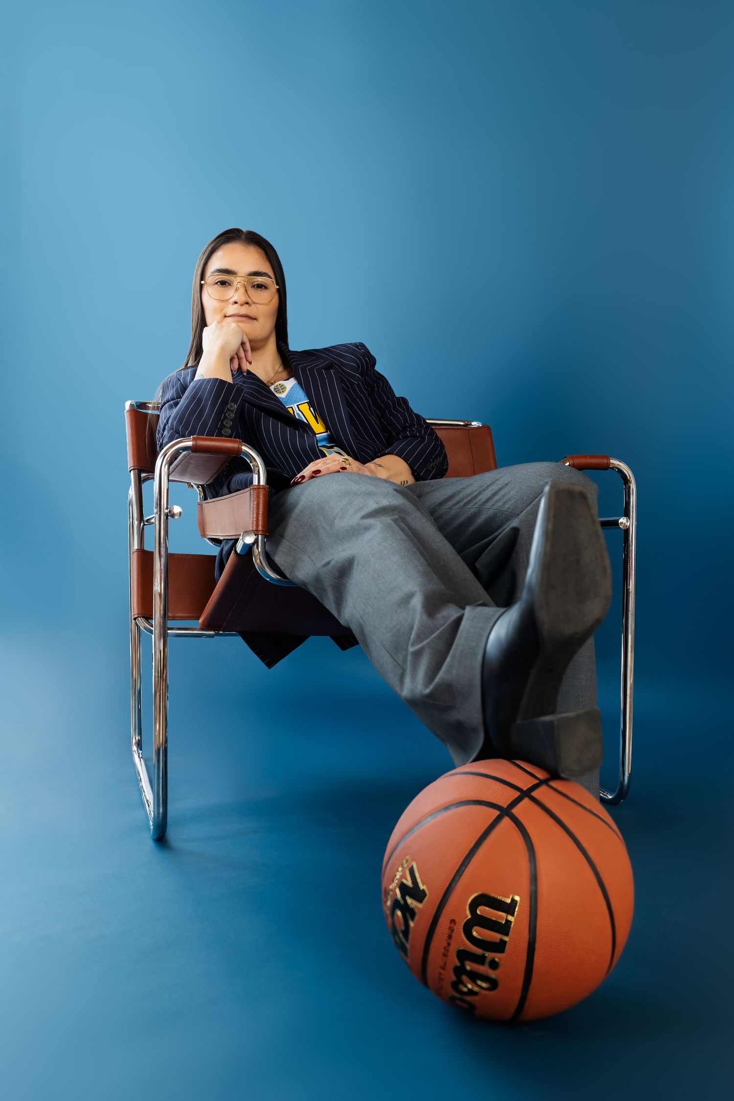 A person in dark clothing reclines across chairs using a basketball as a pillow against a blue studio backdrop.