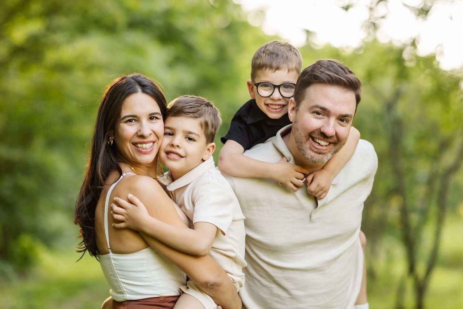 Happy family of four enjoying outdoor time, parents giving piggyback rides to young boys in a green park.