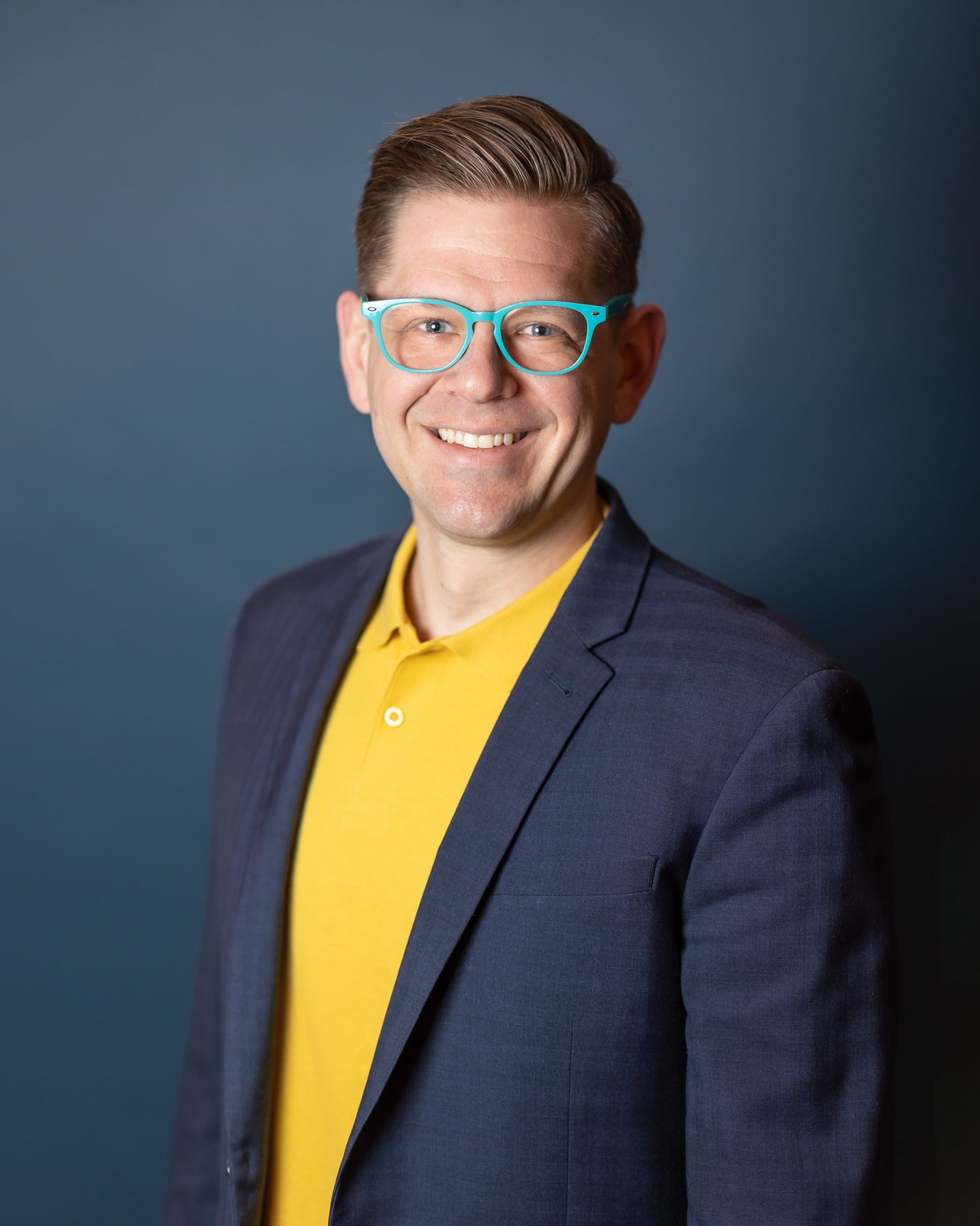 Professional headshot of someone wearing a yellow shirt and navy blazer with blue glasses against dark background.