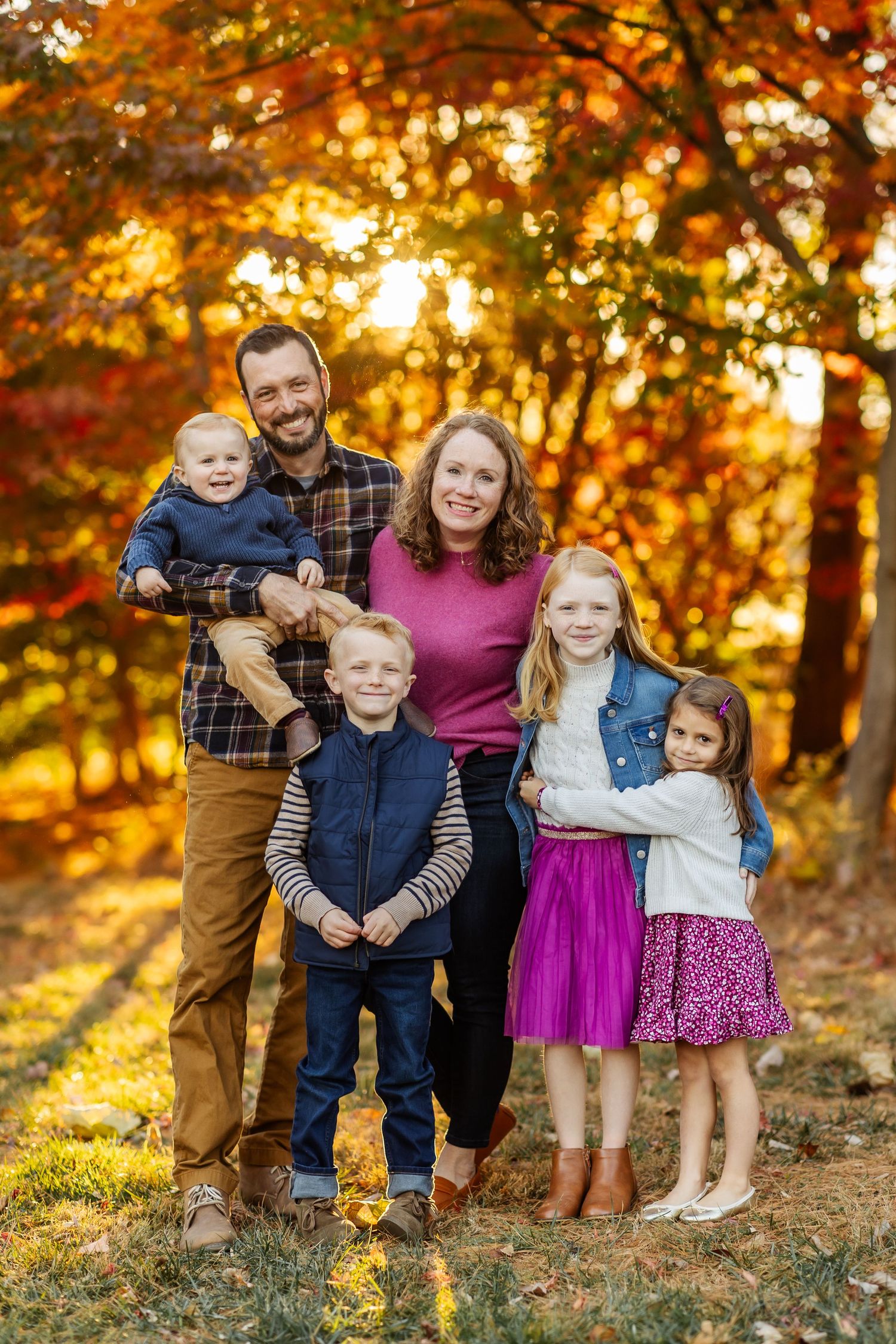 A family portrait captured against vibrant fall foliage with warm sunset lighting.