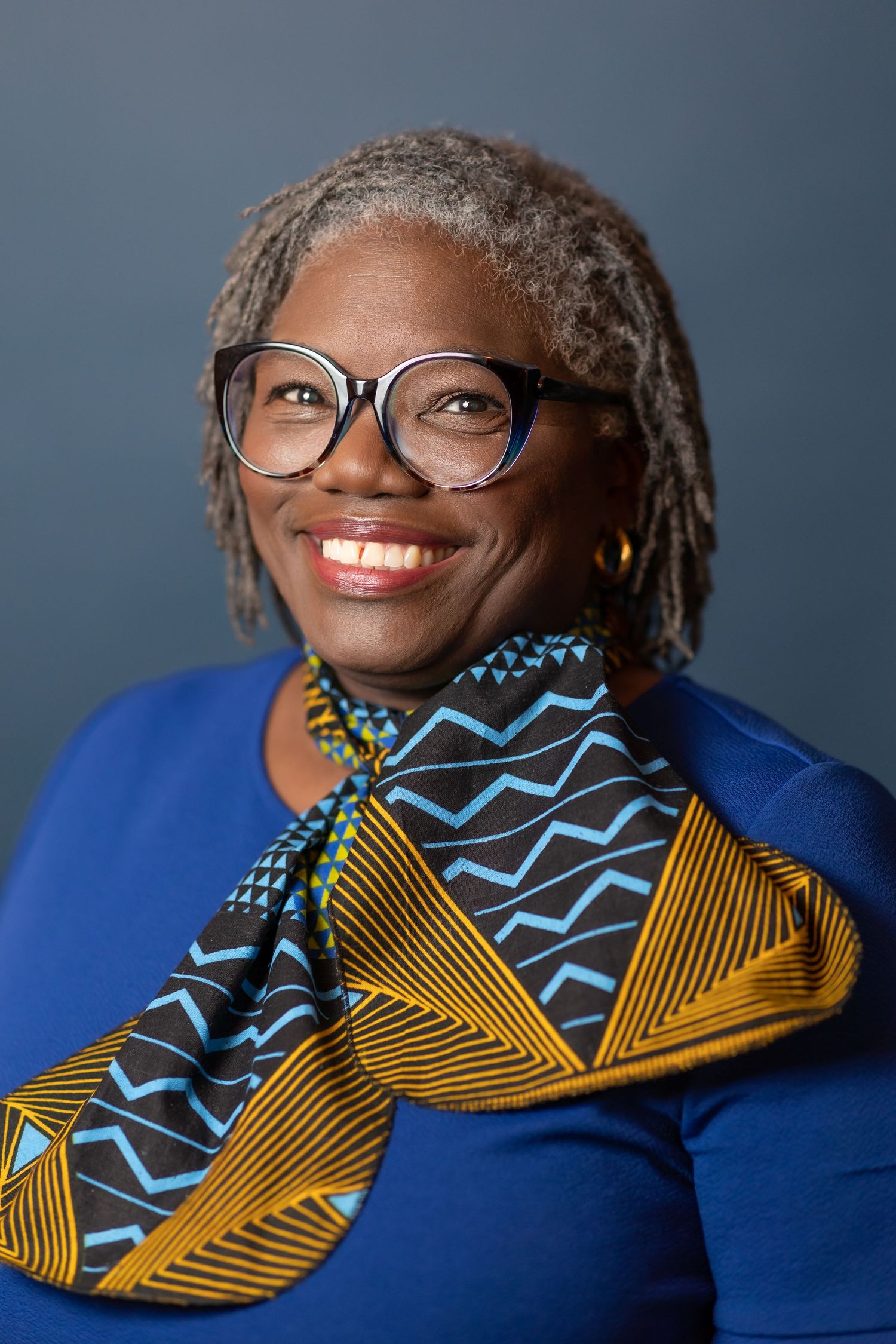 Professional headshot of person in blue top and patterned scarf with geometric designs smiling warmly.
