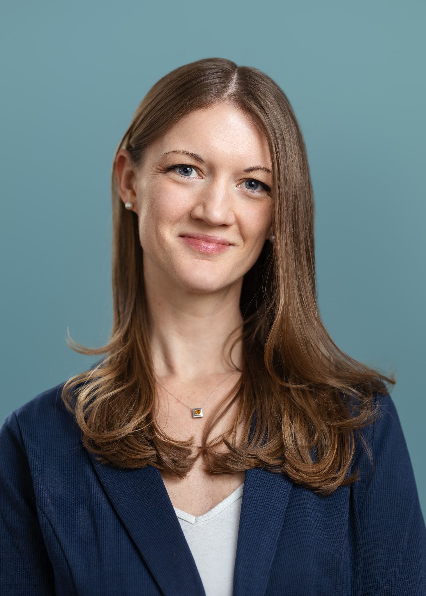 Professional headshot of a smiling woman with brown hair wearing a navy blazer against a teal background.