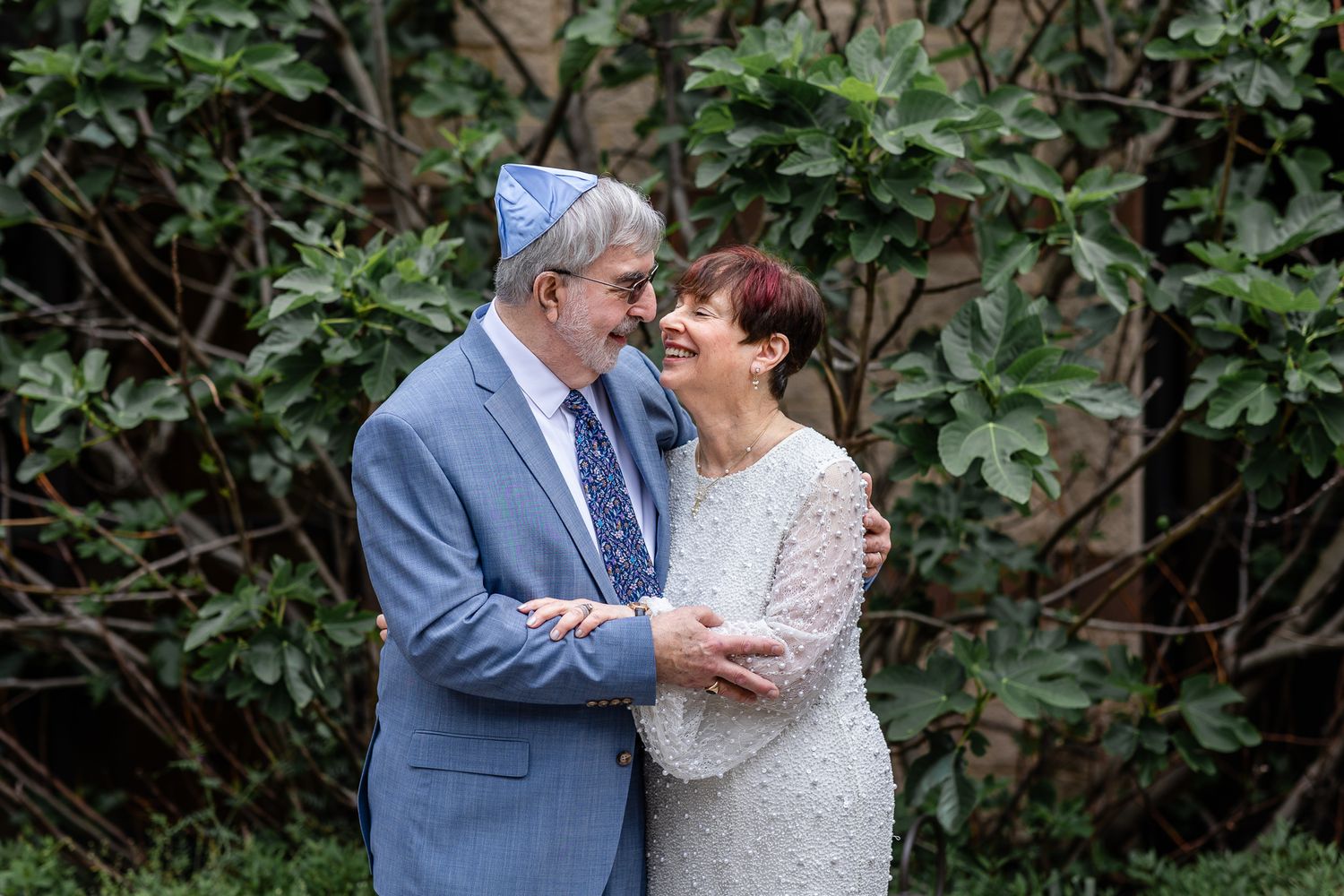 A couple in formal wedding attire embraces in a garden setting with lush greenery in the background.