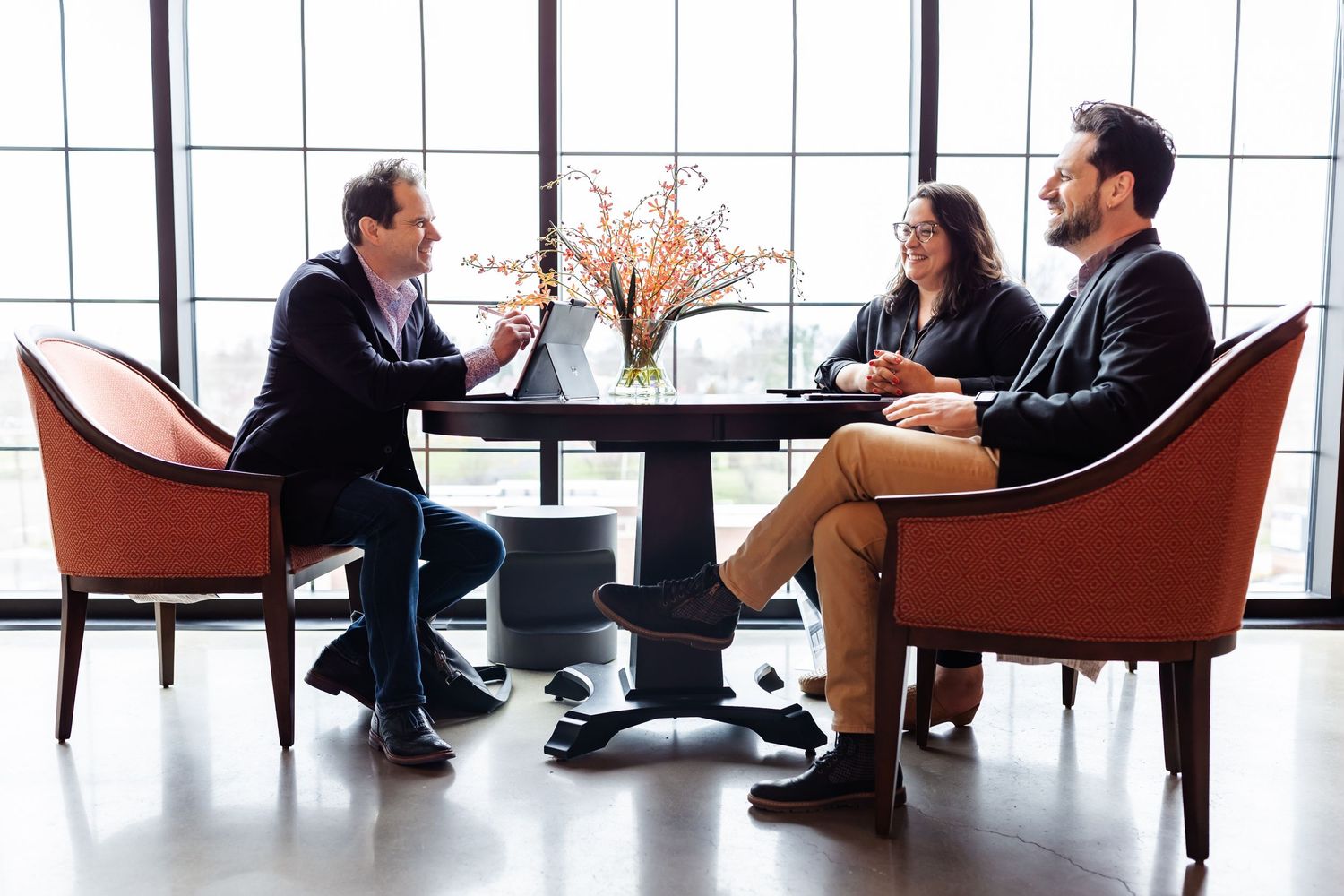 Modern office lounge area with people having casual meetings in orange chairs by large windows.