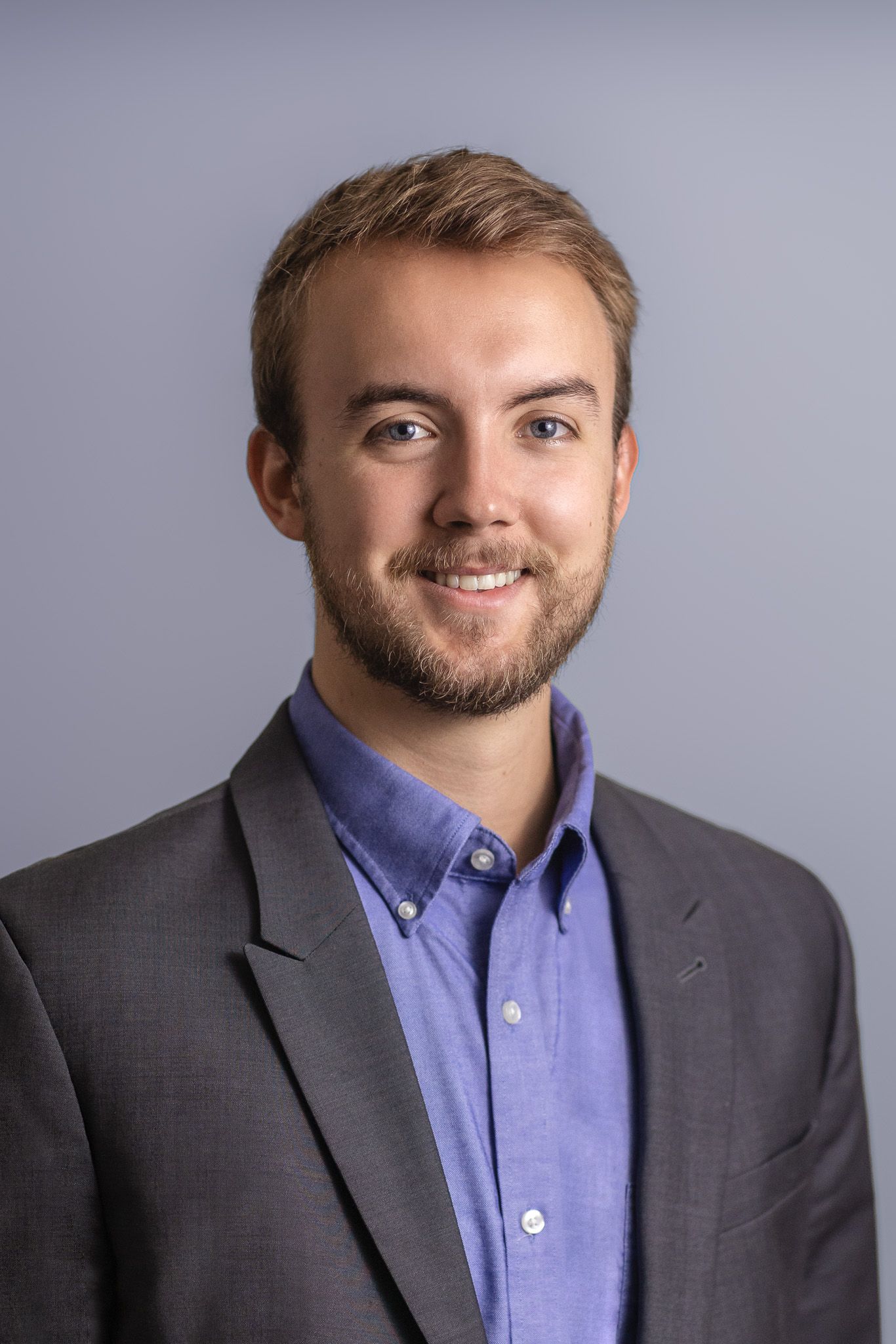Professional headshot of person in grey suit and purple button-down shirt against light grey background.